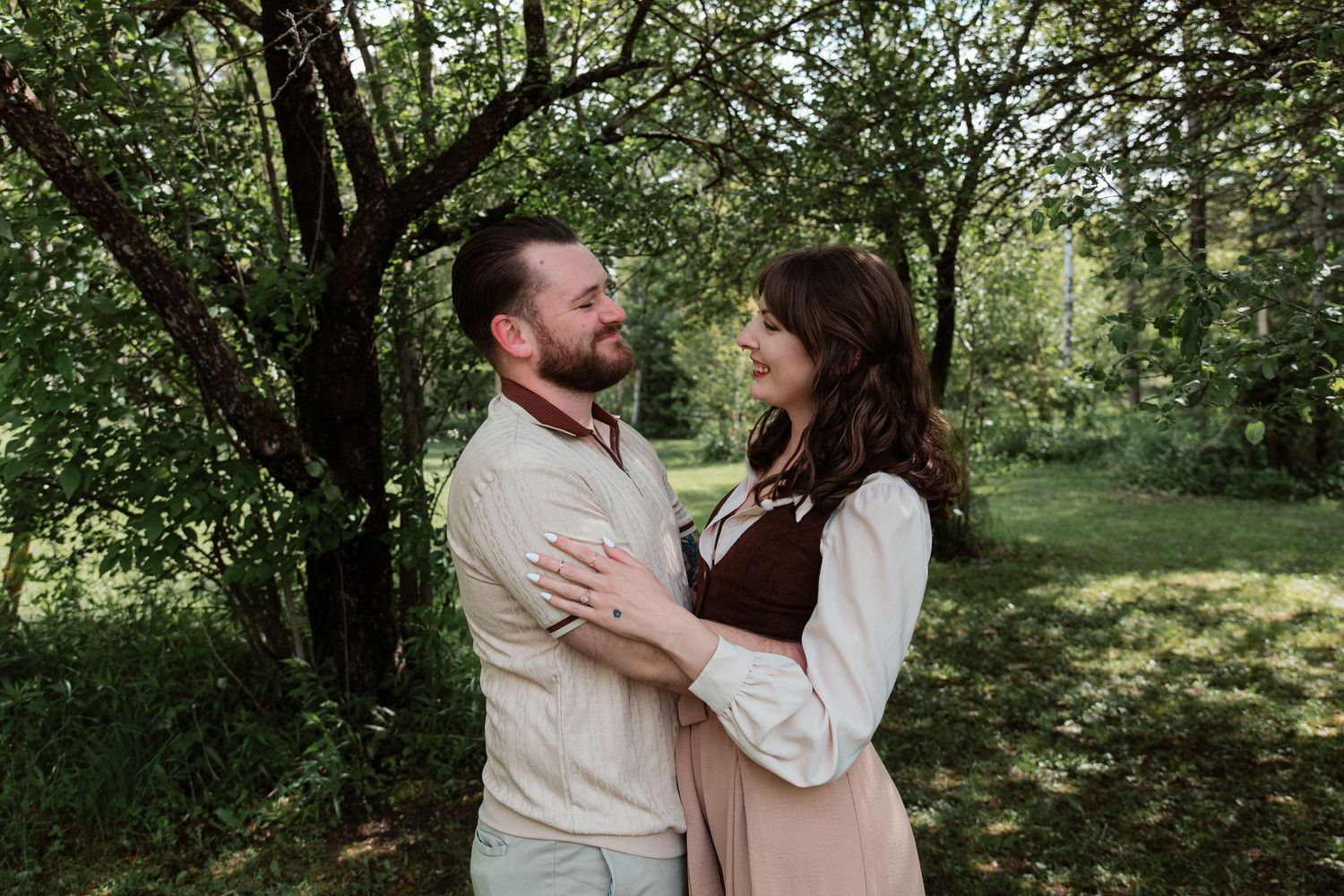 A couple embraces and gazes at each other lovingly outdoors among lush green trees.