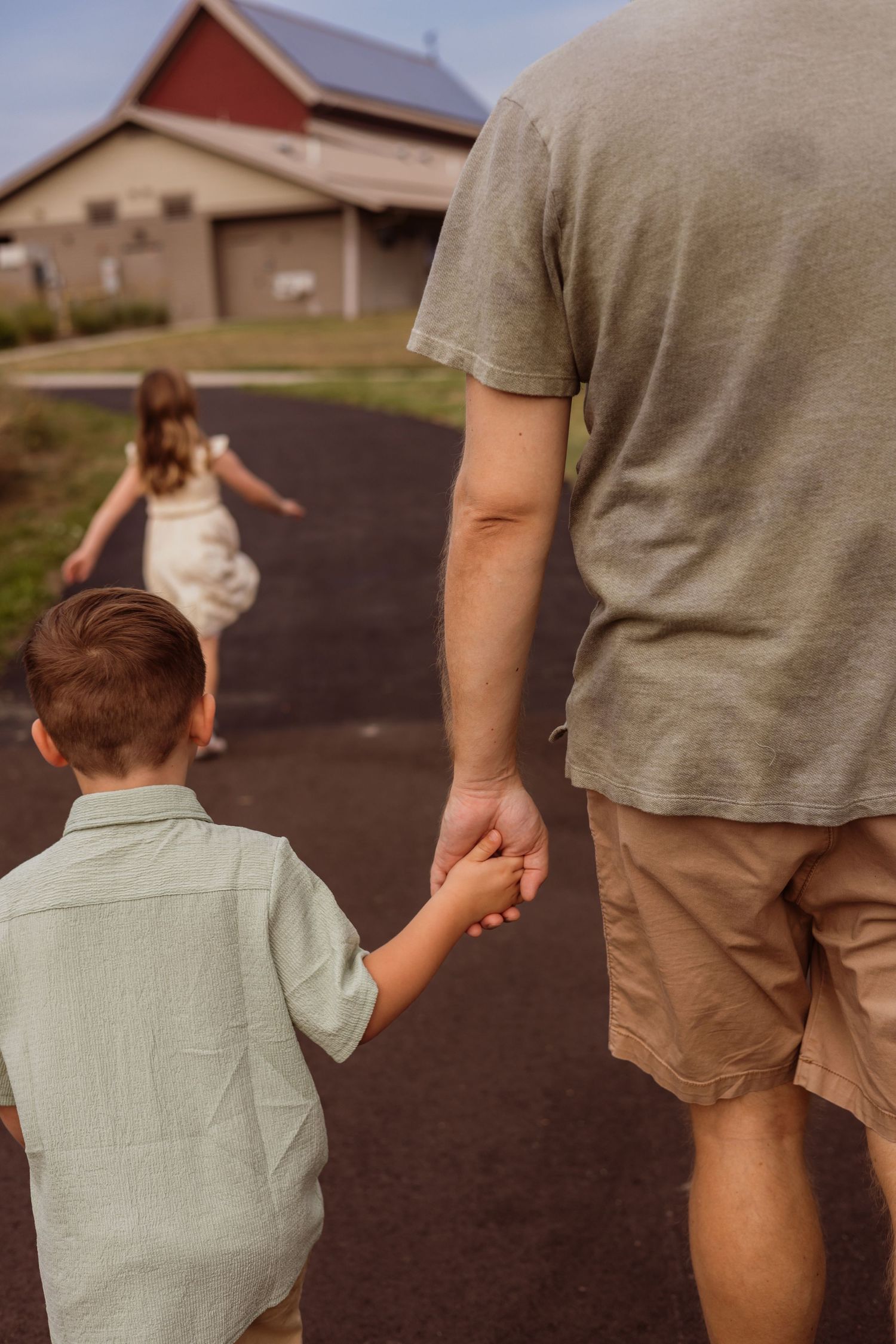 A father holds his young son's hand while walking toward a girl on a rural driveway near a red barn.