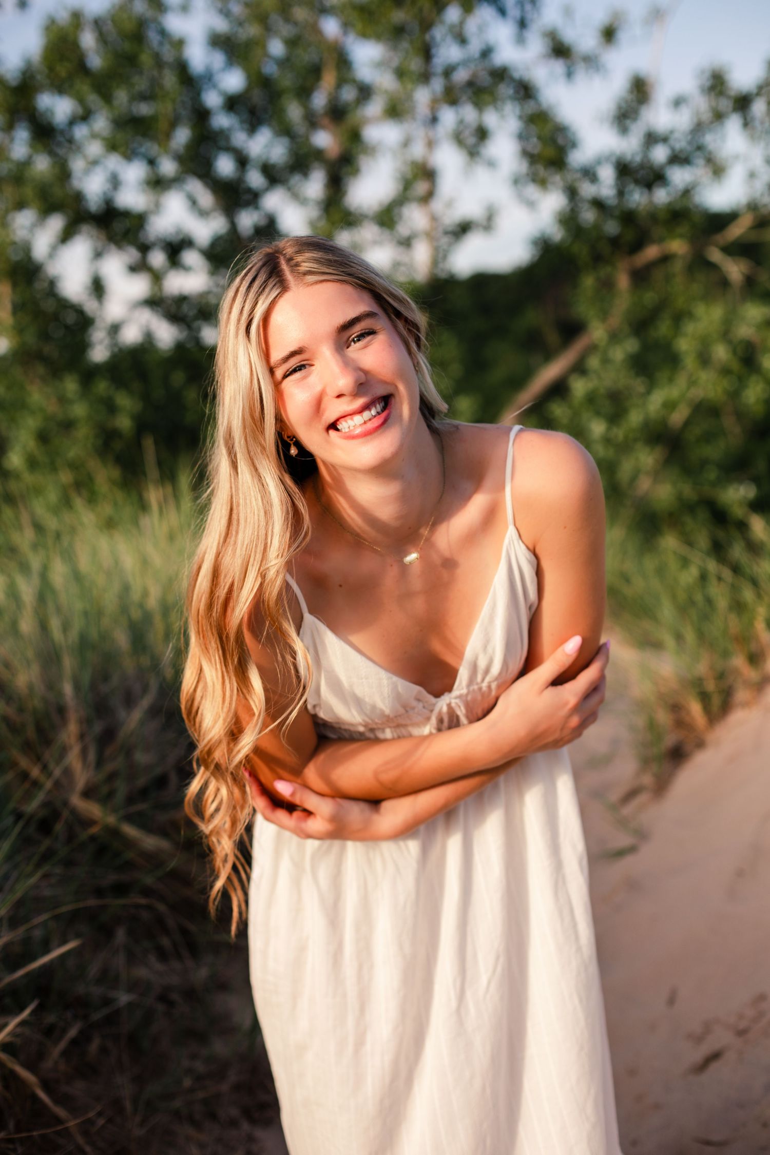 Smiling young woman with long blonde hair wearing a white sundress, standing on a sandy path surrounded by green foliage.