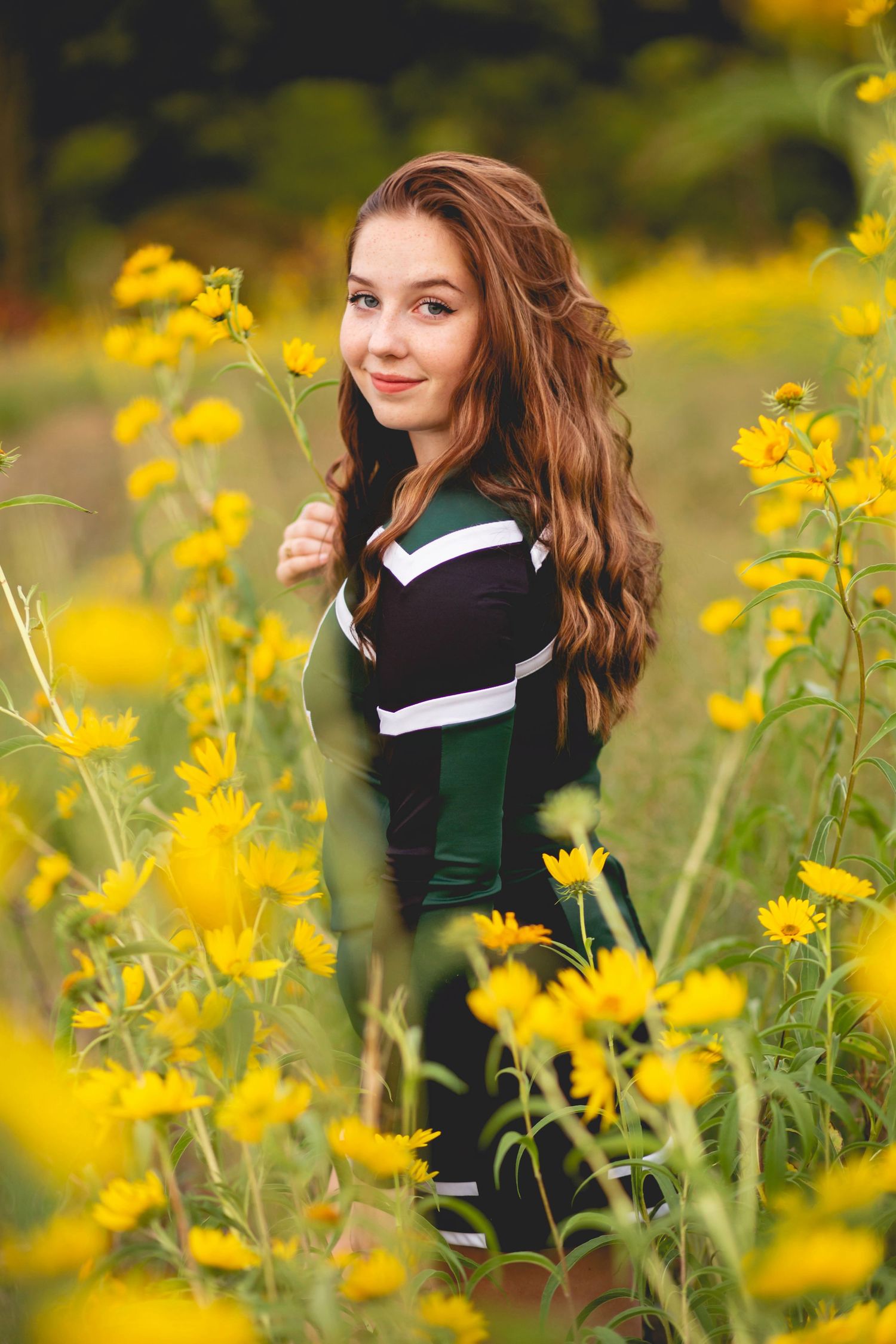 Young woman with long wavy brown hair smiling in a field of bright yellow wildflowers, wearing a green and black outfit.