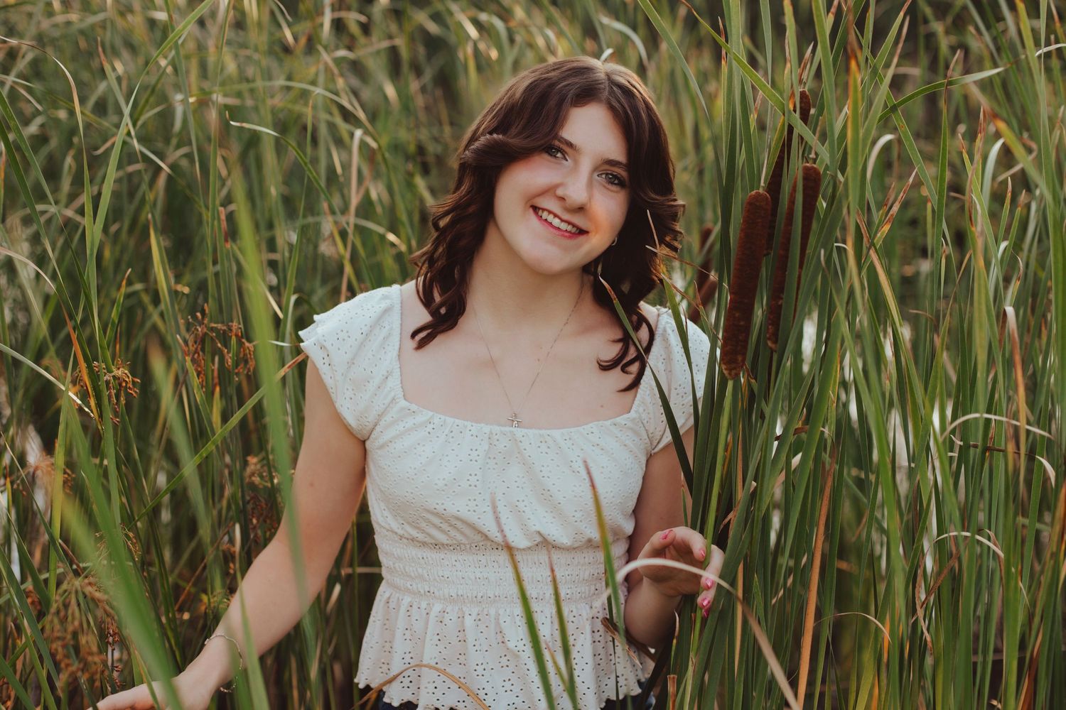 Smiling young woman in white dress posing among tall green cattails in nature.