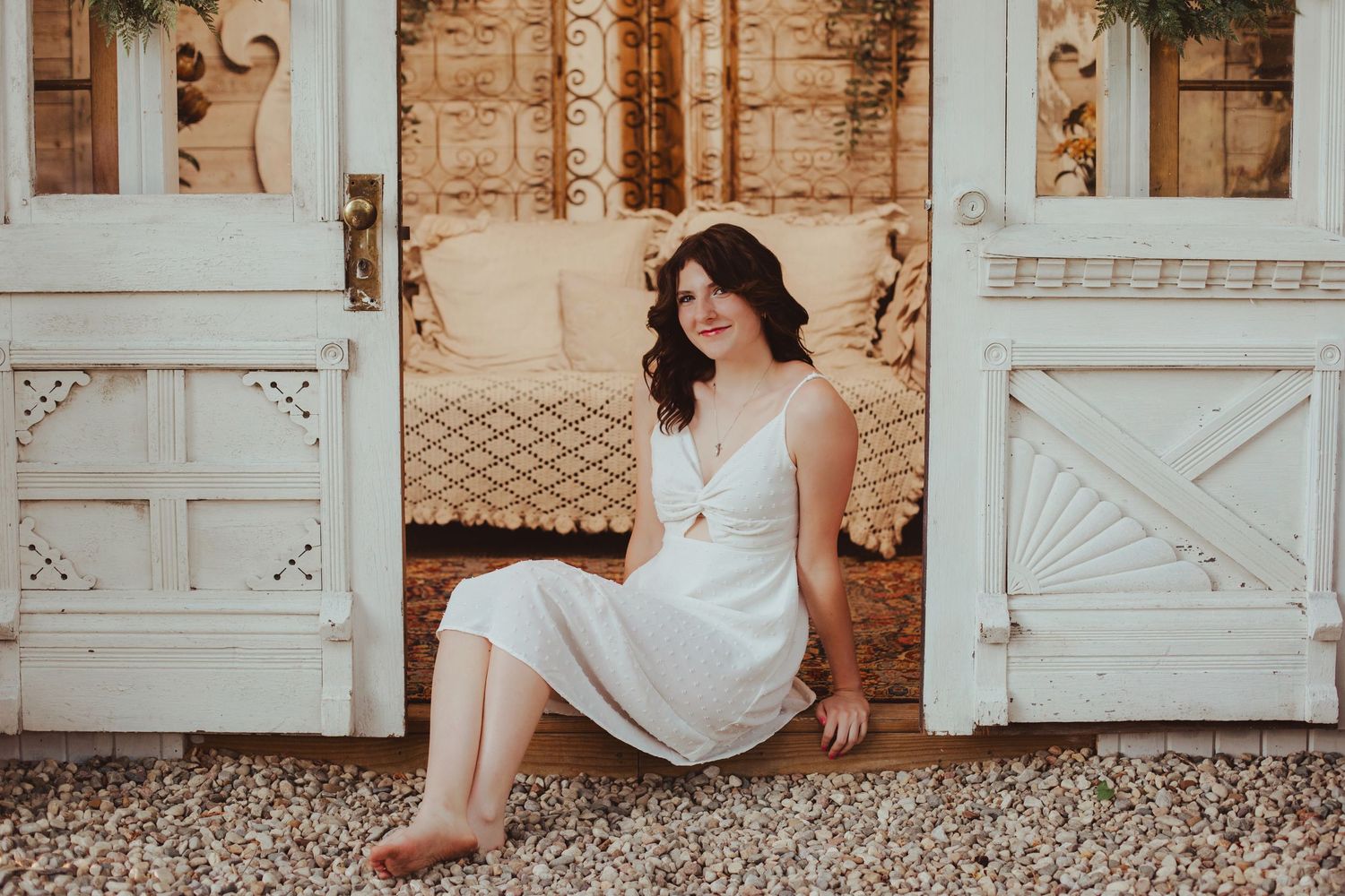 Young woman in white dress sitting barefoot in front of vintage white doors.