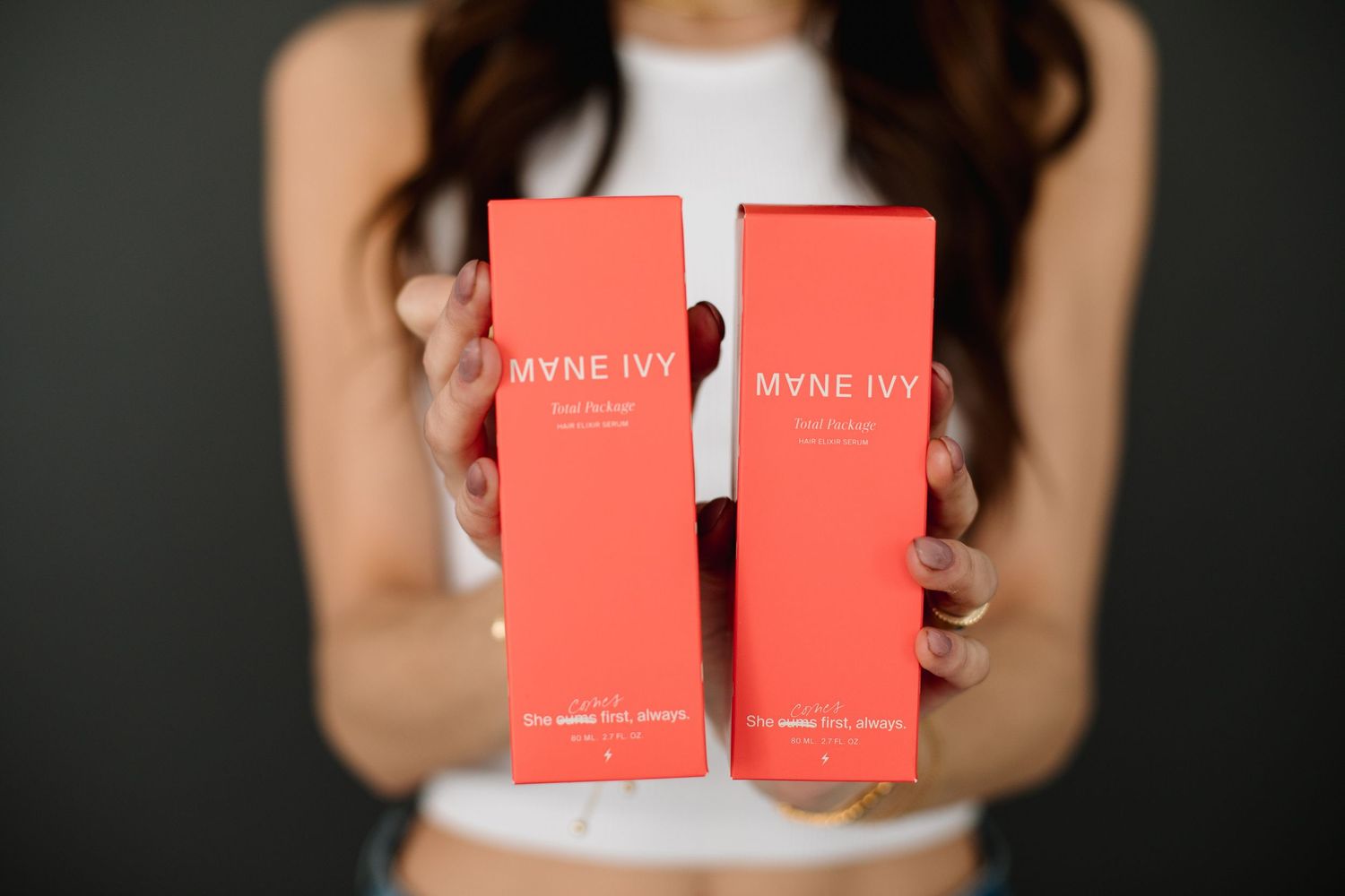 Woman holding two coral Mane Ivy hair product boxes against a dark background.