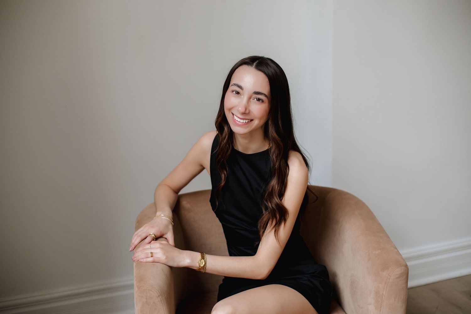 Smiling brunette woman seated in a velvet chair wearing a black dress with a gold bracelet.