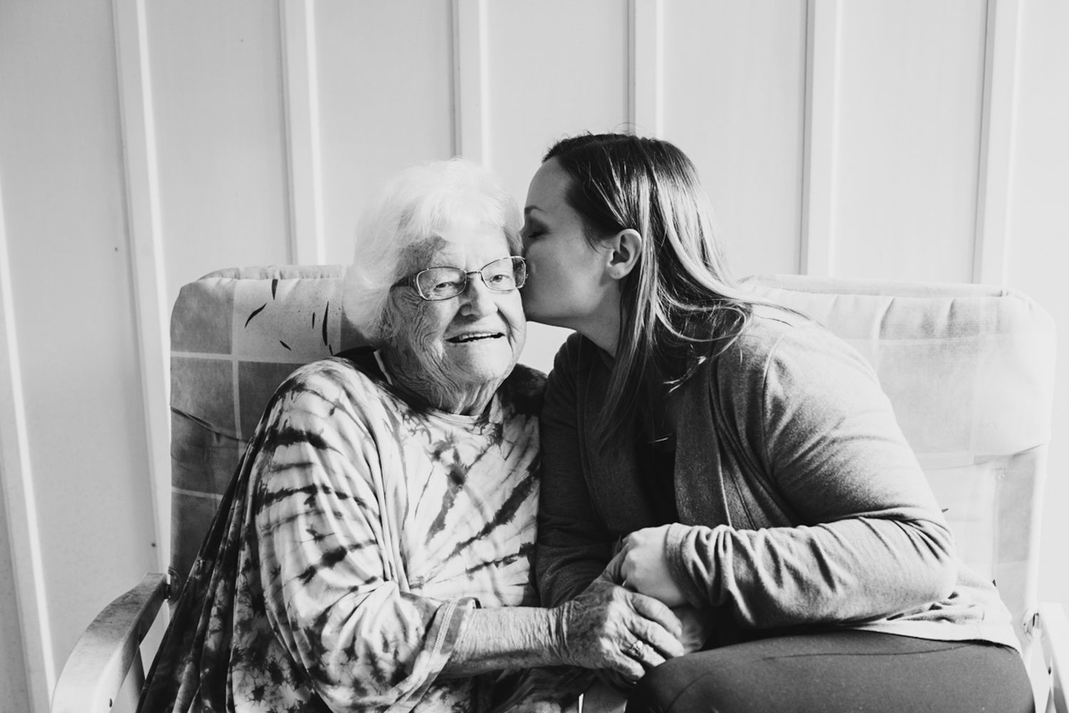 Young woman kissing elderly grandmother on cheek while sitting together in black and white photo.