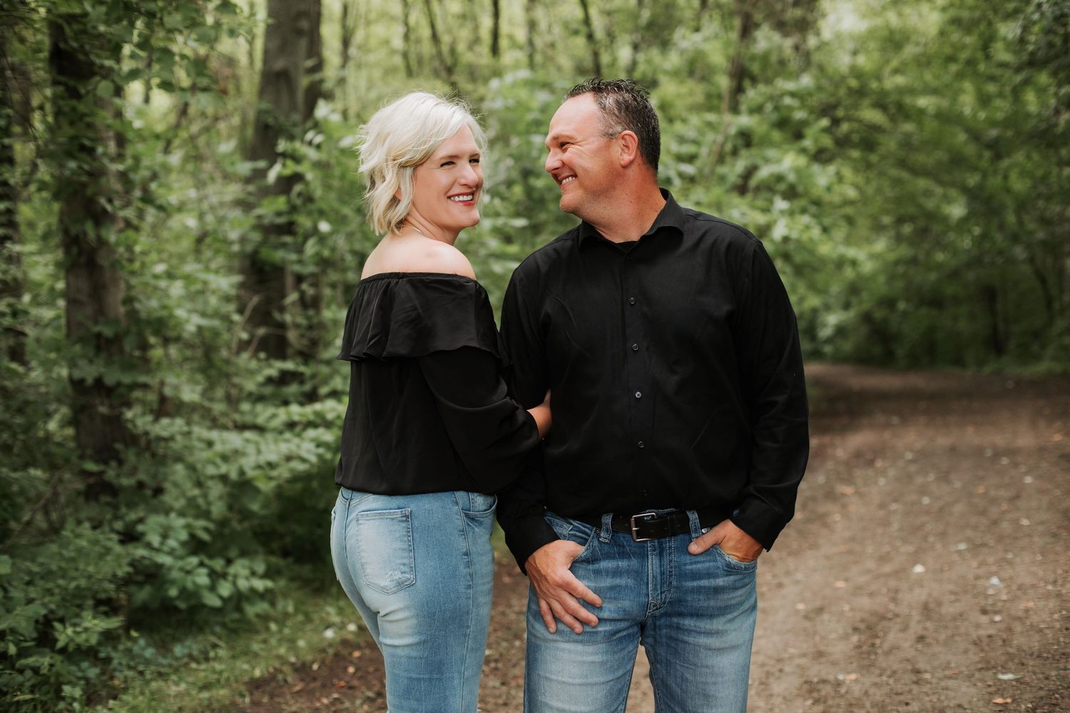 A cheerful couple in black tops walk hand in hand along a tree-lined dirt path in the woods.