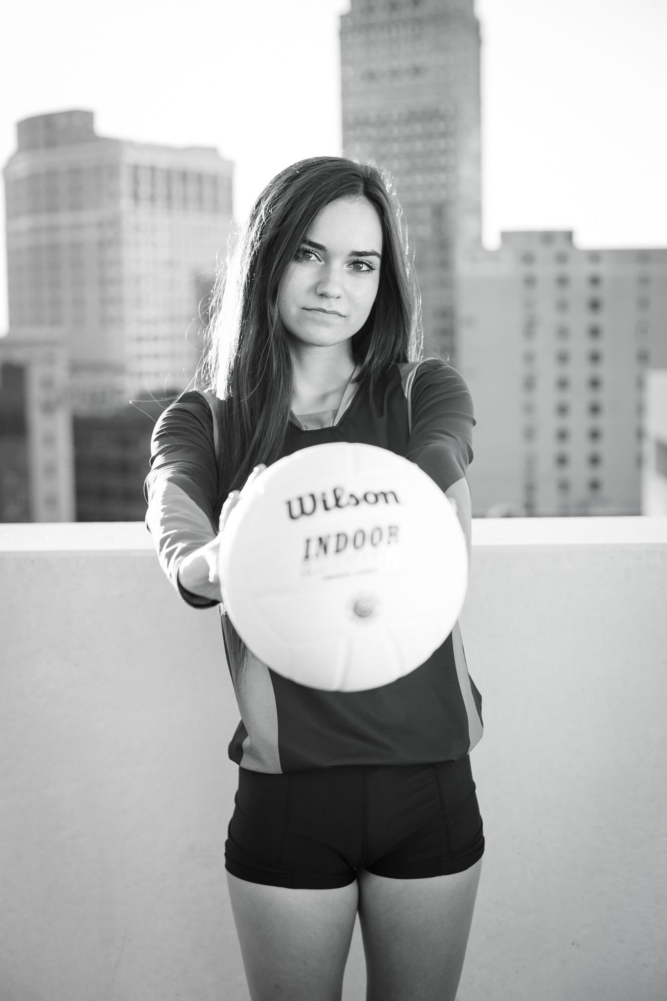 Young female athlete holds Wilson indoor volleyball toward camera on urban rooftop in black and white.