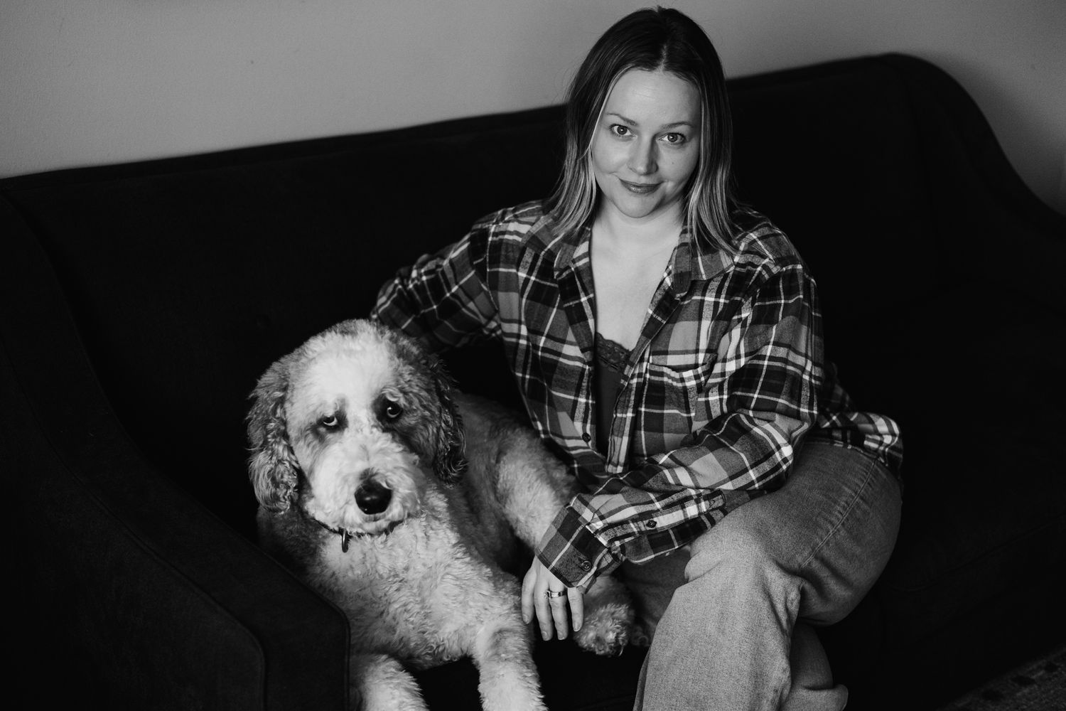Black and white portrait of a smiling woman sitting on a sofa with a fluffy white doodle dog.