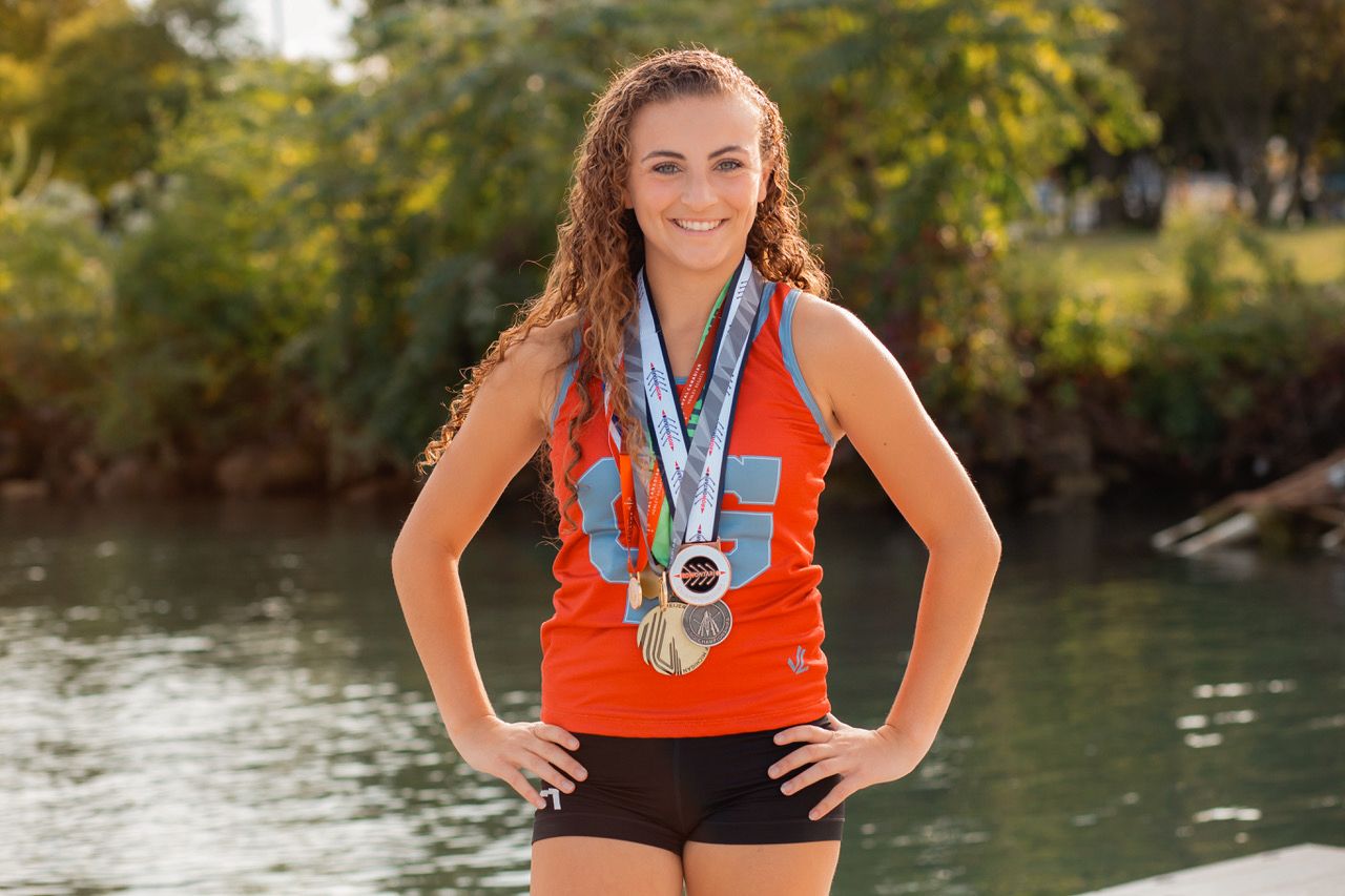 Smiling young athlete in orange jersey proudly wears multiple medals near a riverside backdrop.