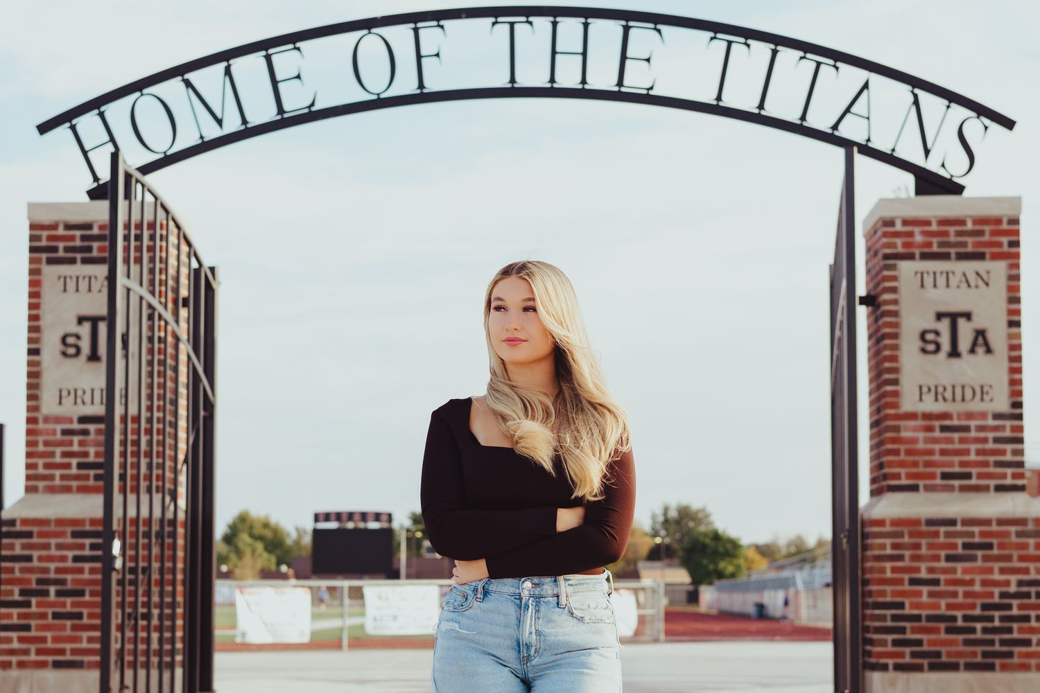 Blonde teenage girl crosses arms under Home of the Titans arch for high school senior portrait session.