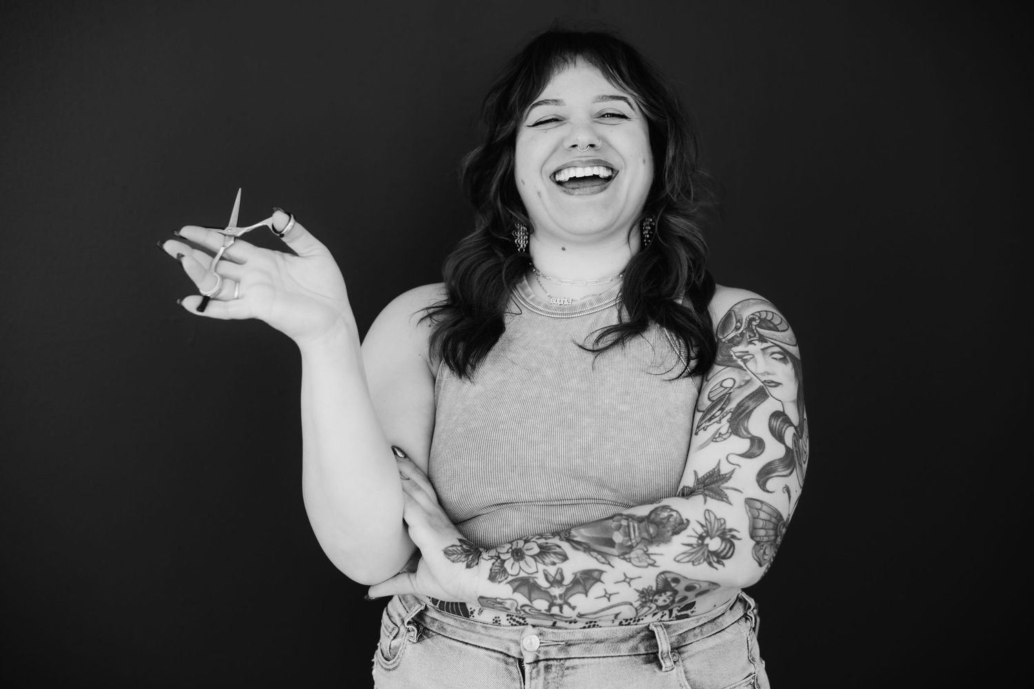 A laughing woman with tattoos holds scissors while posing against a dark background in black and white.