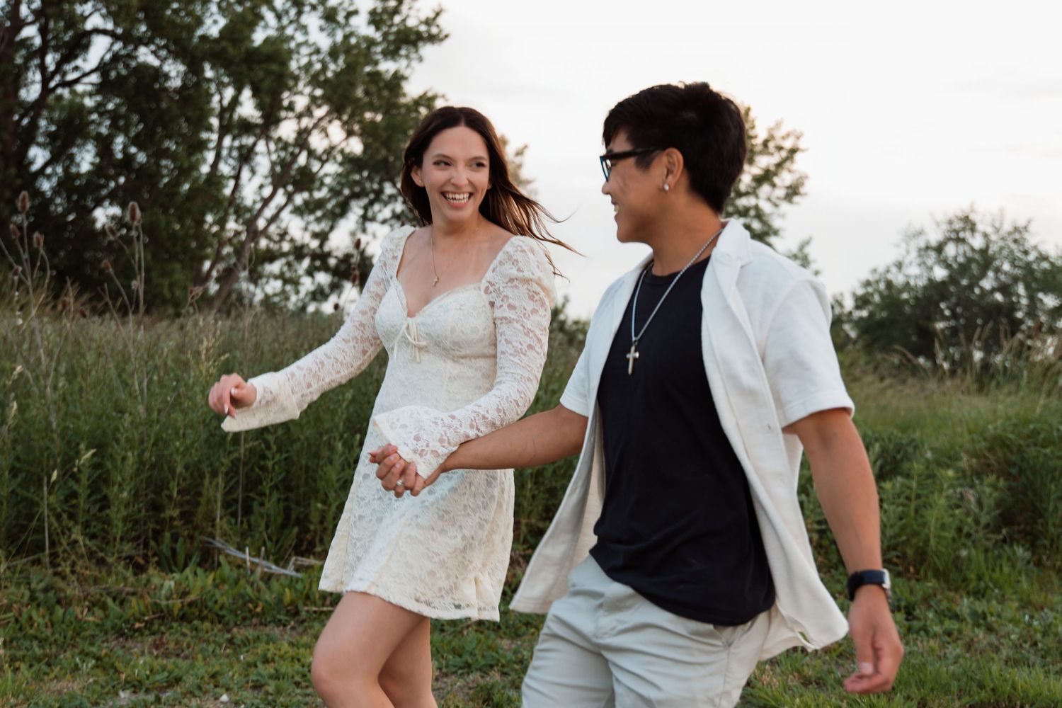 A smiling couple walks hand in hand through a lush green field during golden hour sunset light.