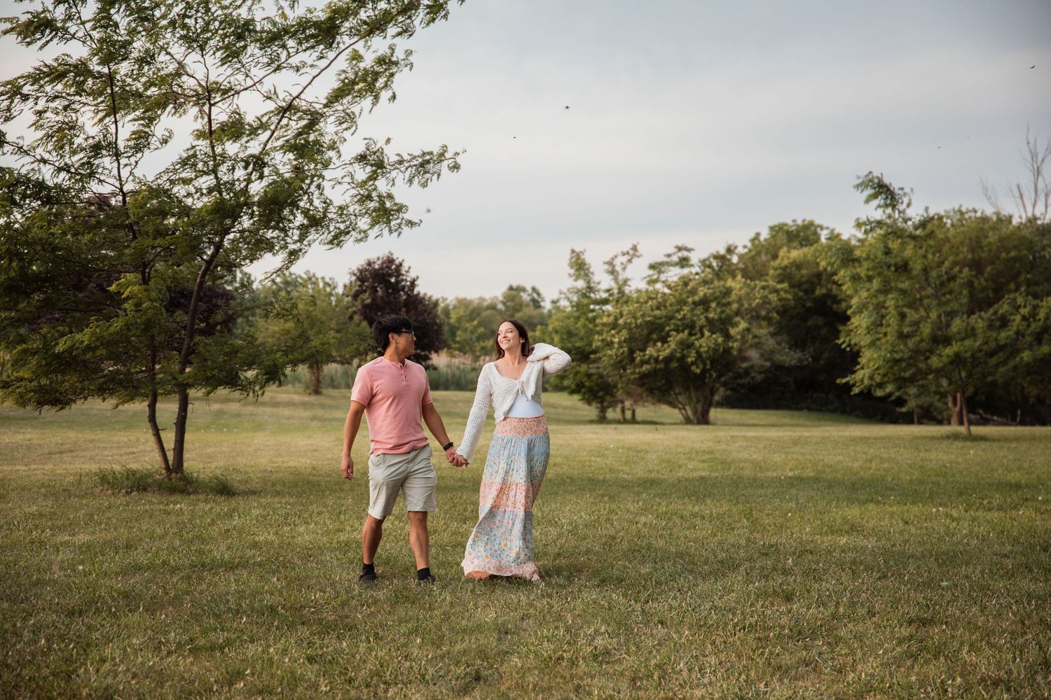Couple holding hands walking in a lush green park with trees in the background on a cloudy day.