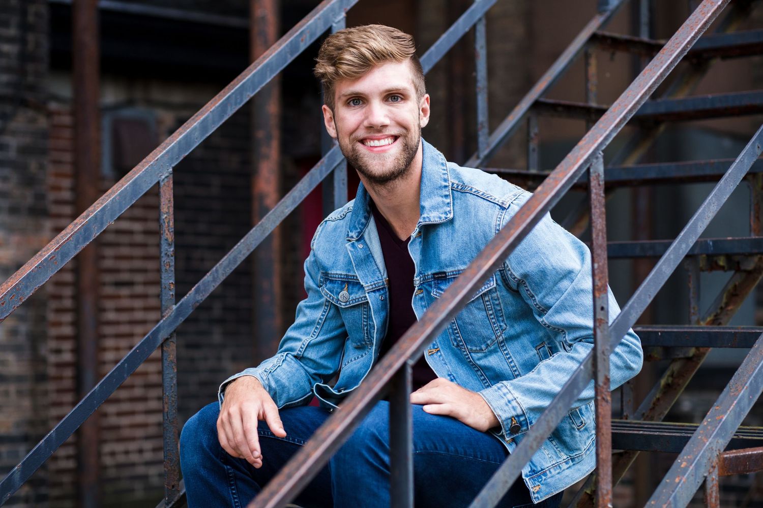 Young man in denim jacket smiling on urban metal fire escape stairs outdoors.