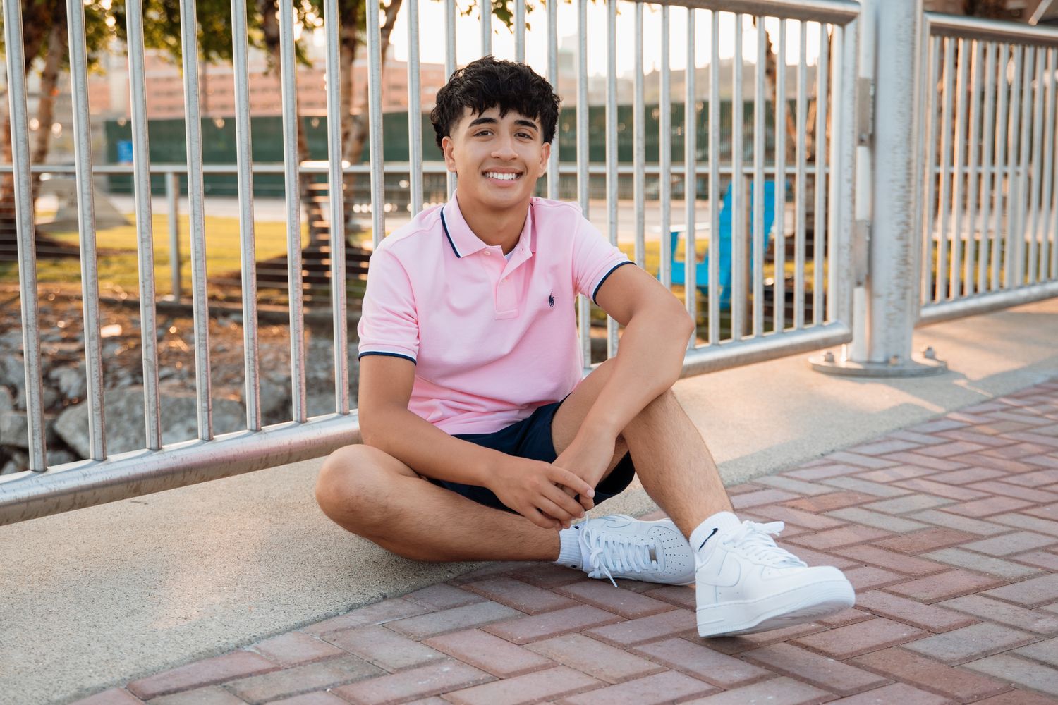 Smiling young man in pink polo shirt and shorts sitting on pavement near metal railing outdoors.