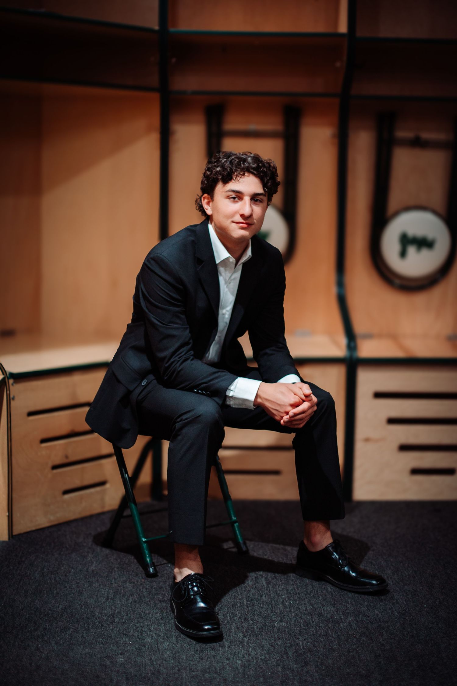 Young man in black suit sitting confidently on chair in modern wooden interior setting.