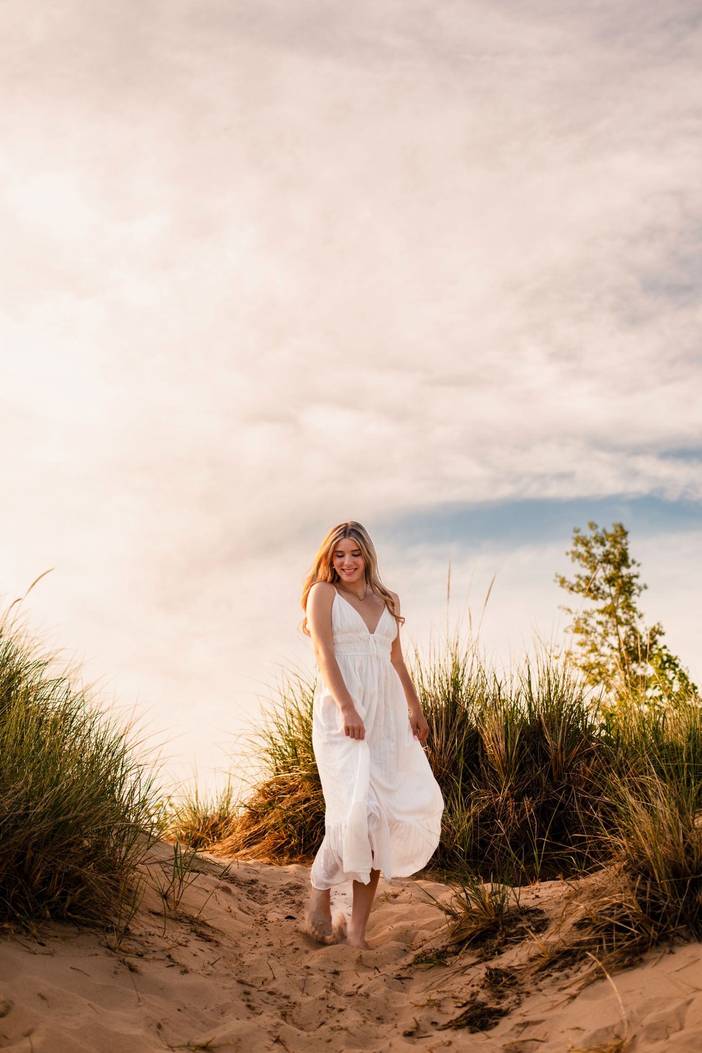 Smiling blonde woman in white maxi dress standing on sandy dunes with green grass and cloudy sky.