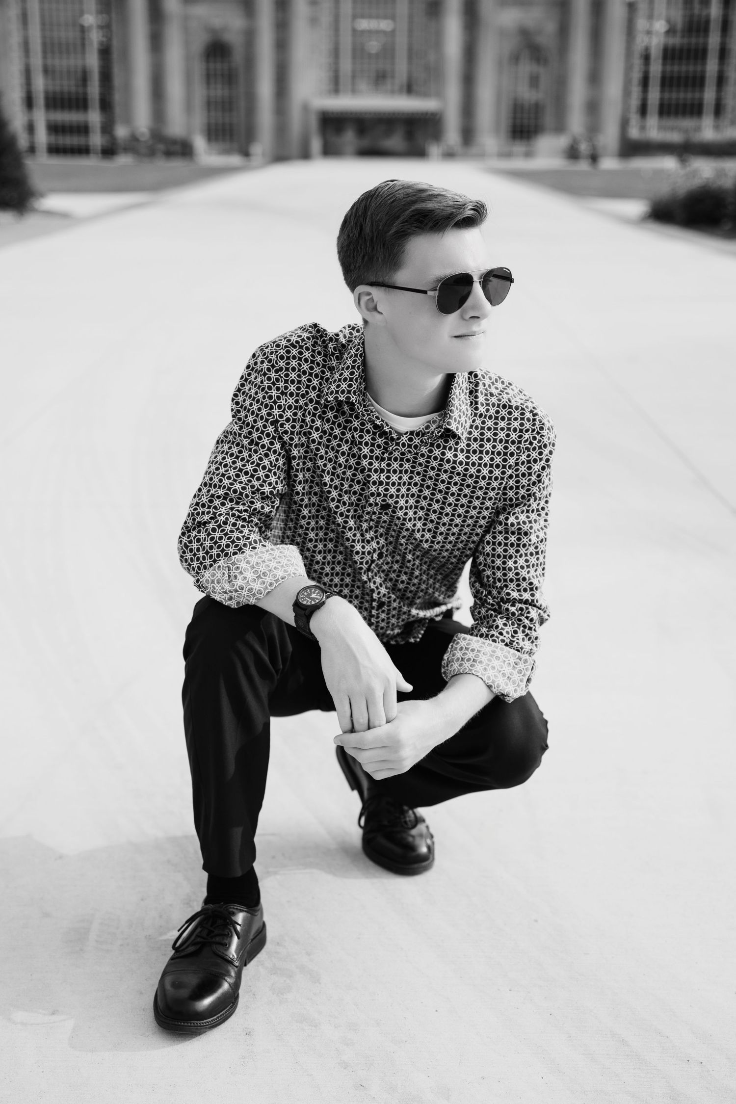 Black and white portrait of a young man in sunglasses and patterned shirt crouching outdoors.