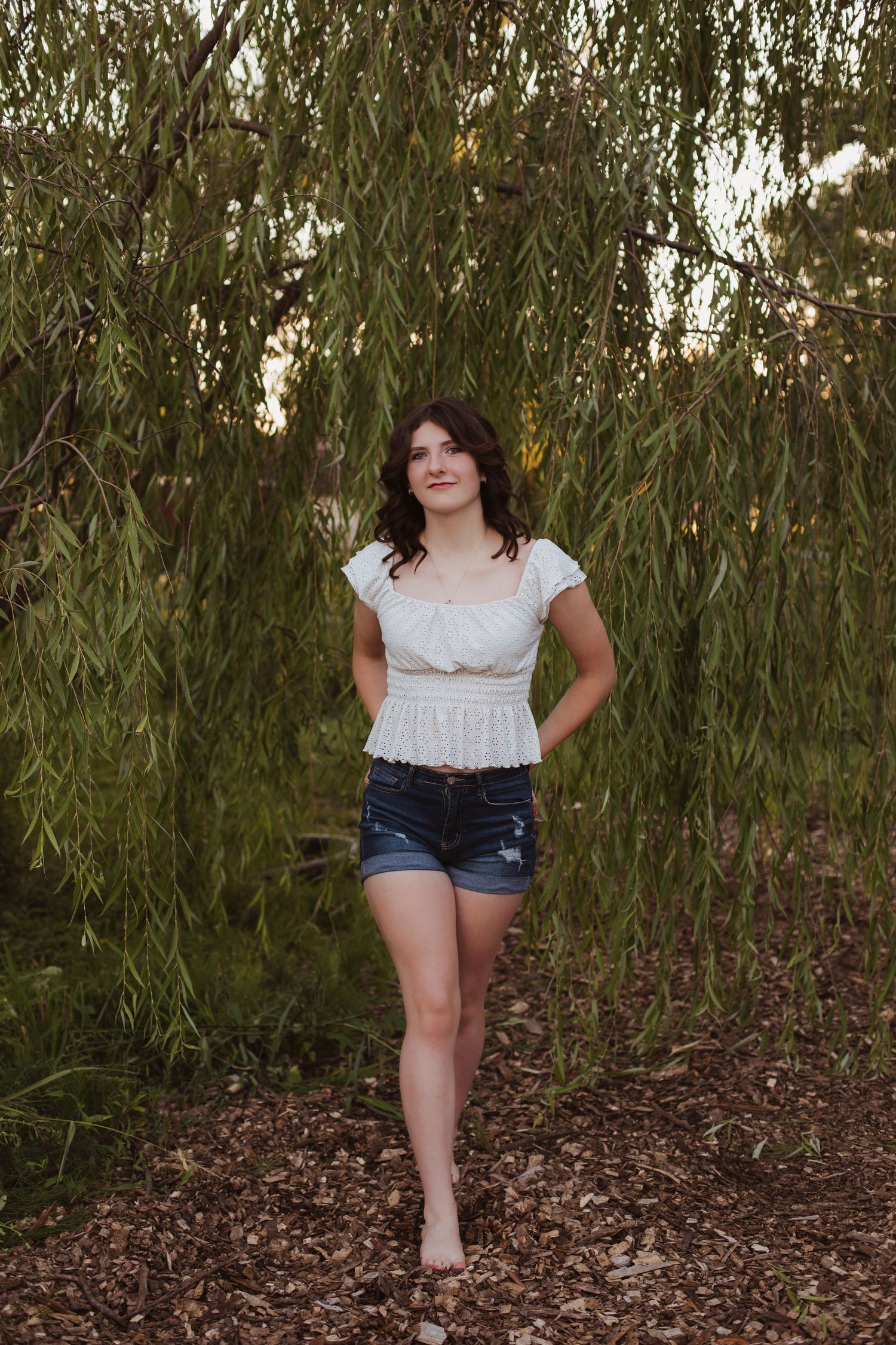 Young woman in white top and denim shorts standing under a weeping willow tree.