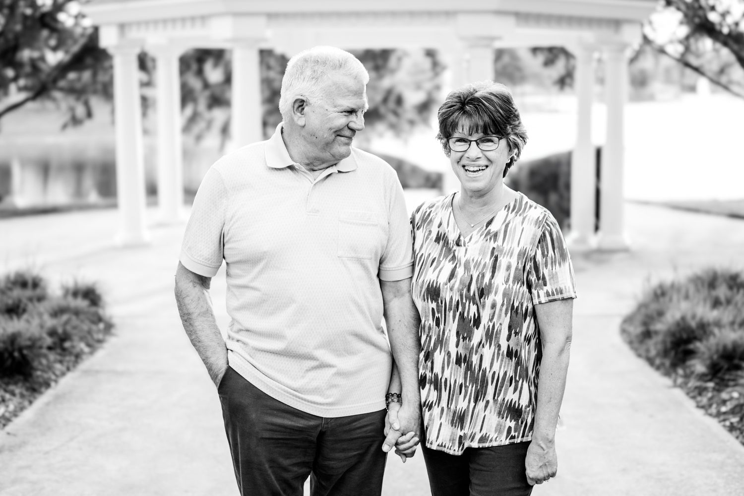 A smiling couple holds hands on a garden path with a gazebo backdrop in a black and white photo.
