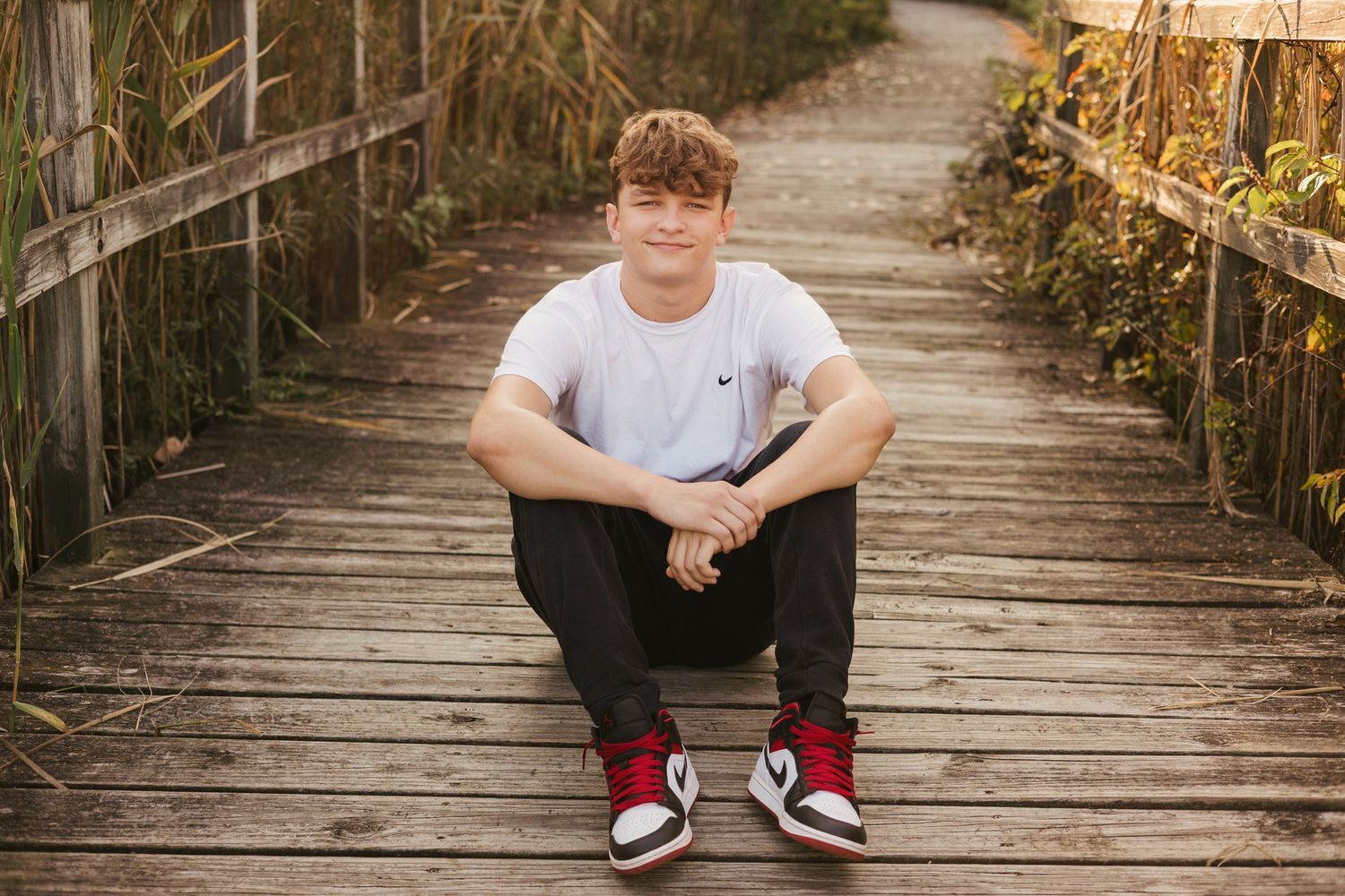 Teen boy in white Nike tee and red Air Jordan 1s sits on wooden boardwalk path surrounded by tall grass.