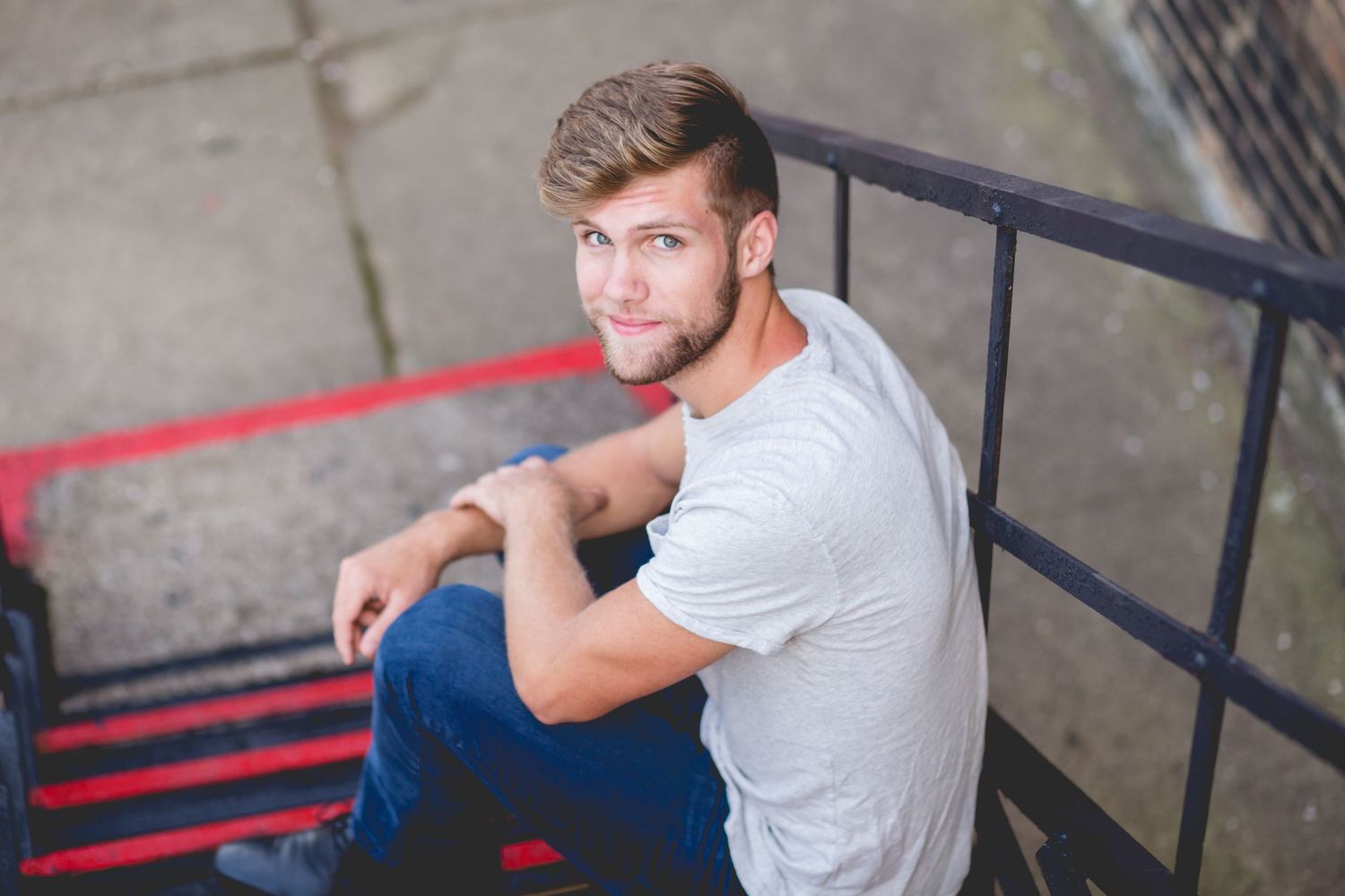 Young man with beard sitting on red metal stairs outdoors, wearing white t-shirt and jeans, smiling at camera.