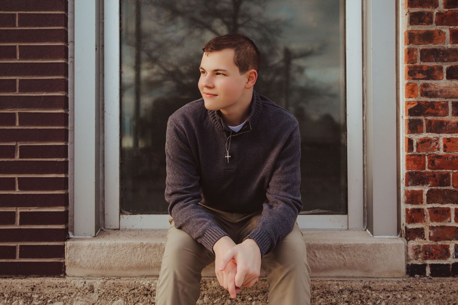 Young man in gray sweater with cross necklace sits on brick steps, gazing thoughtfully to the side.