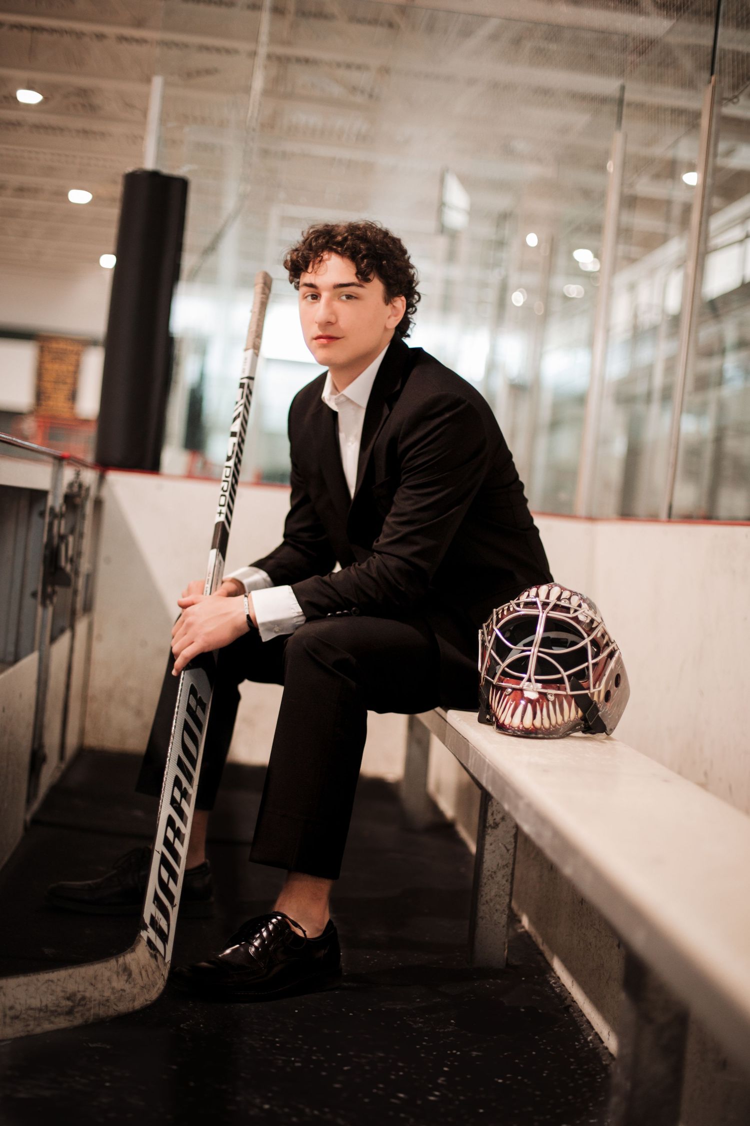Young hockey goalie in black suit holds goalie stick beside helmet on rink bench in professional portrait.