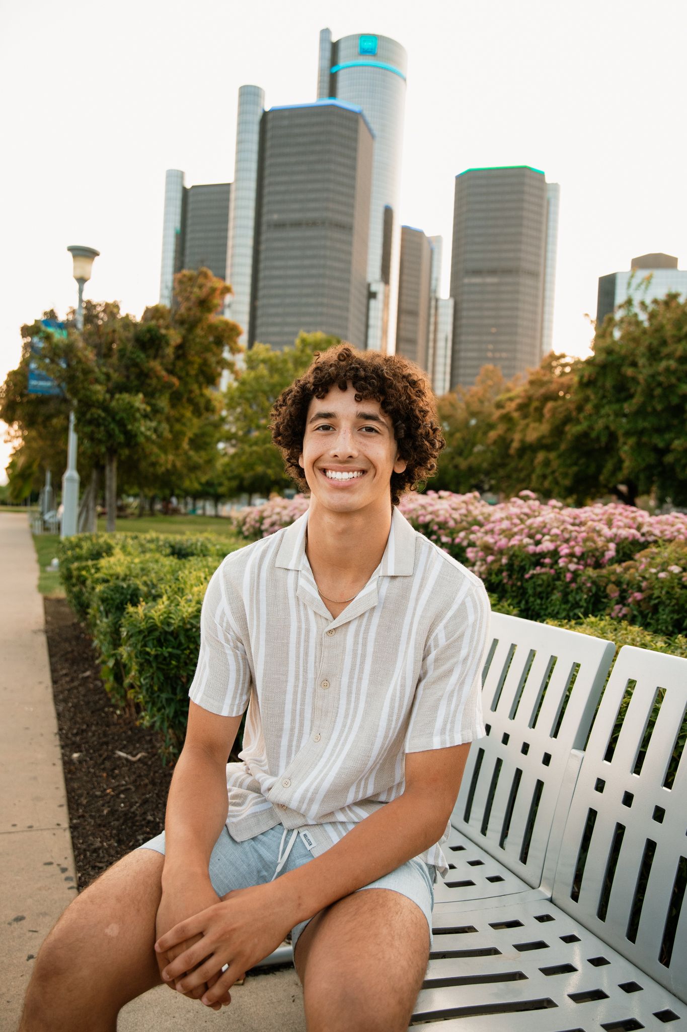 Young man with curly hair smiling on a white bench in a city park with skyscrapers in the background.