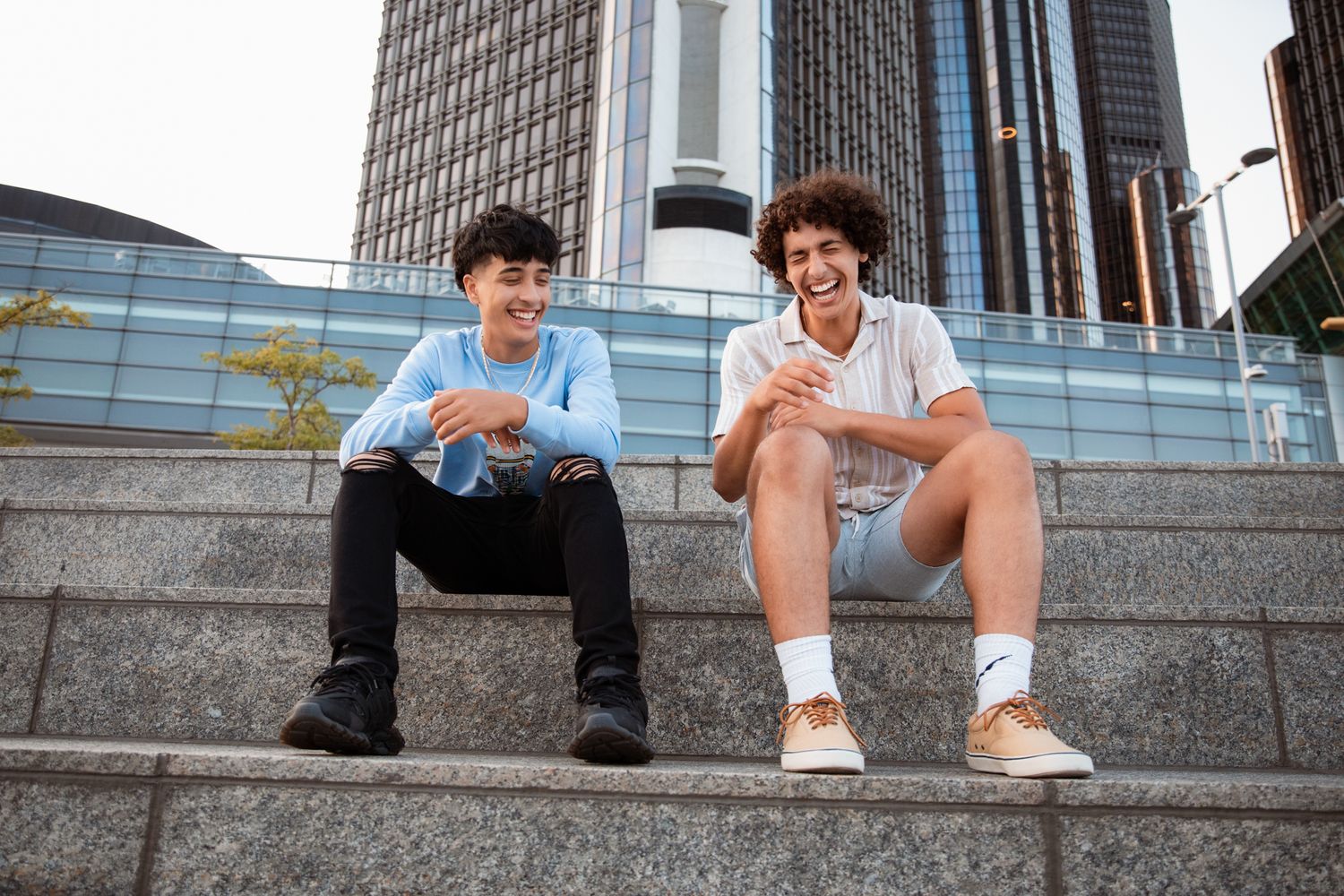 Two young men sitting on outdoor steps laughing and using smartphones with a modern skyscraper in the background.