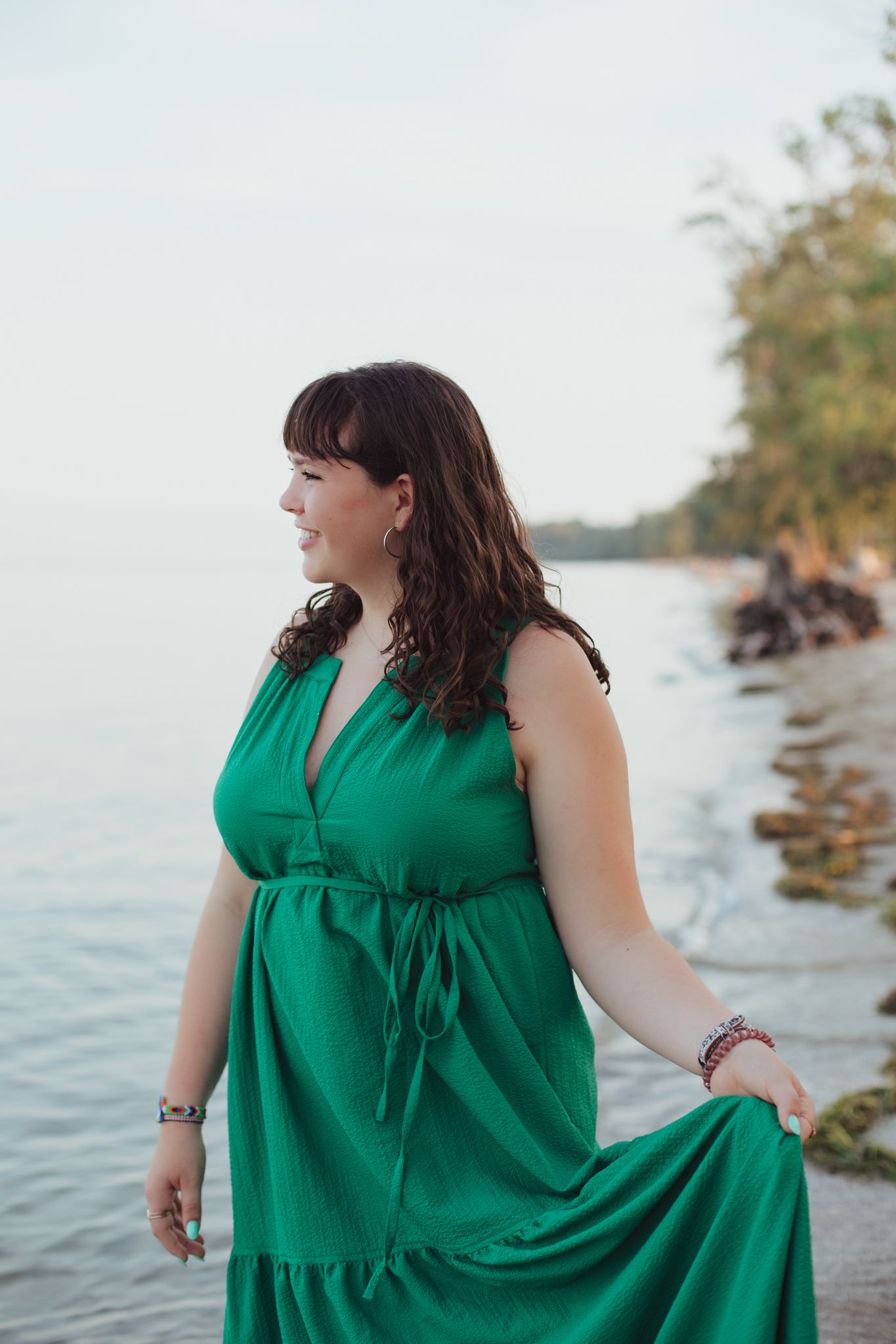 Pregnant woman in green maxi dress standing on rocky shoreline, smiling sideways with trees and calm water behind her.