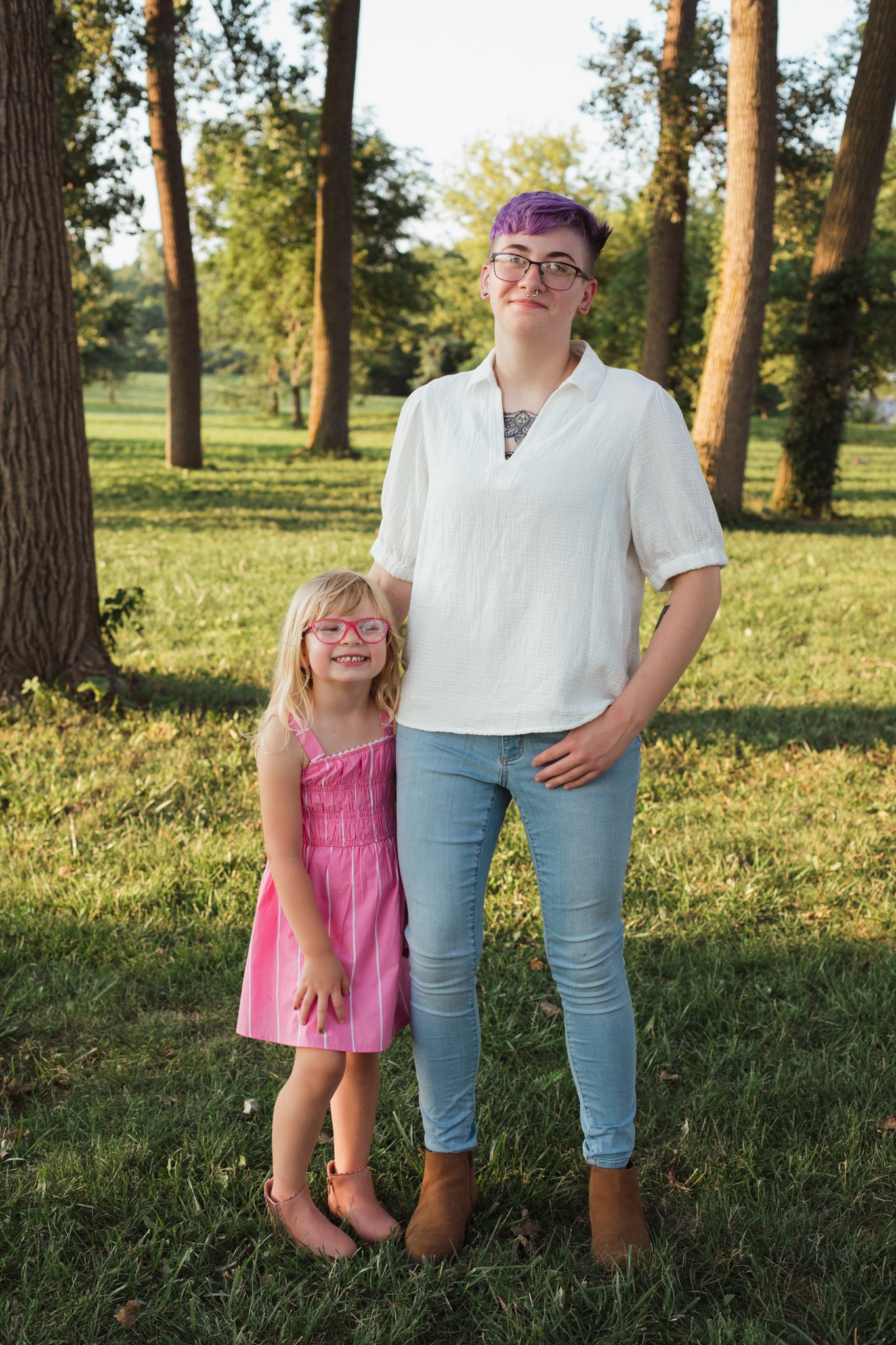Two girls posing outdoors in a sunny park, one in a pink dress with glasses, the other in white top and jeans.
