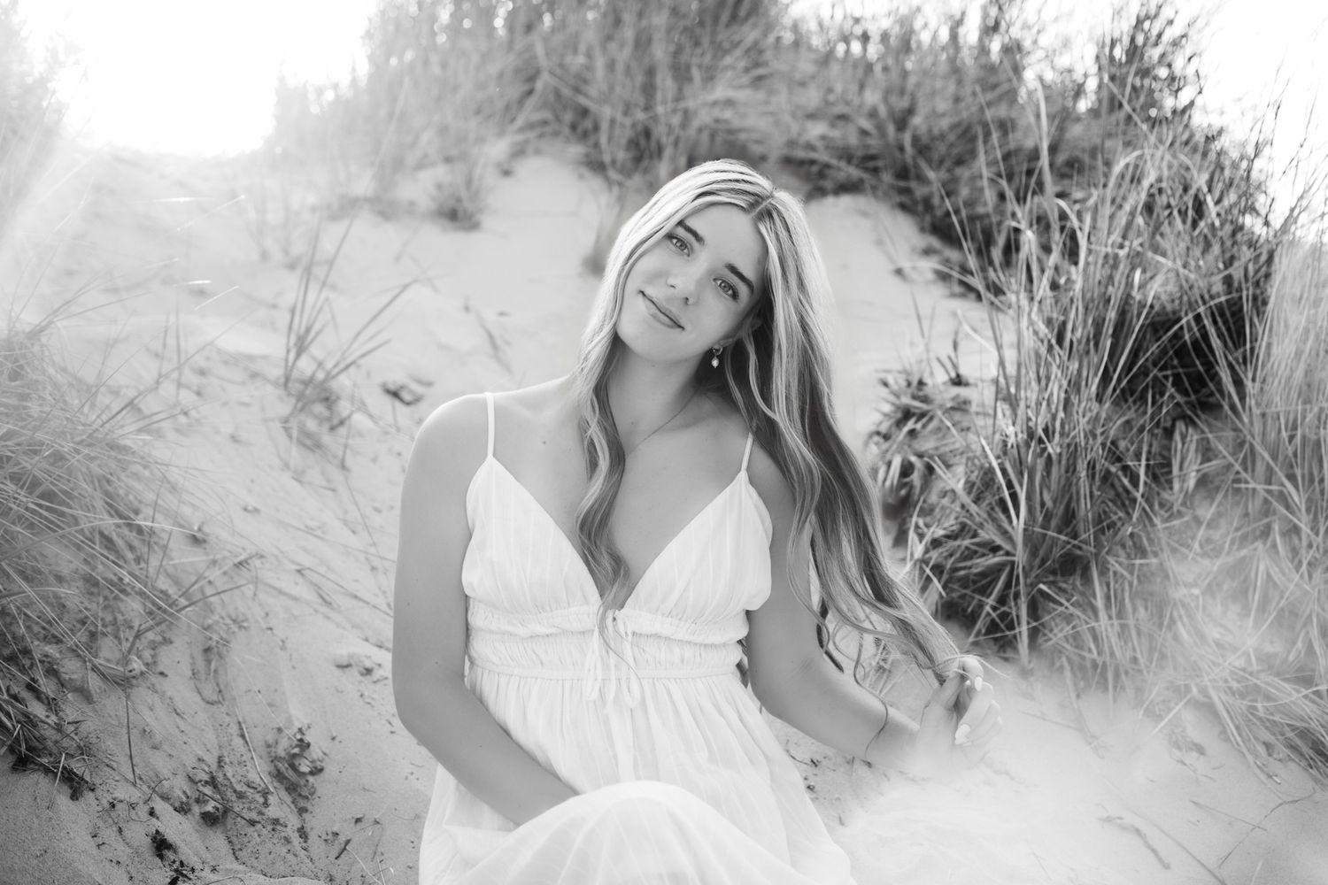 Black and white photo of a young woman in a white dress sitting on sandy dunes surrounded by tall grass.