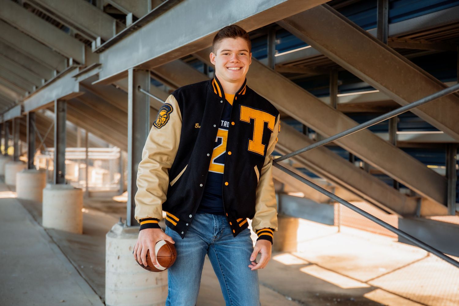 Teen boy in varsity letterman jacket holding football, smiling under stadium bleachers in senior portrait.