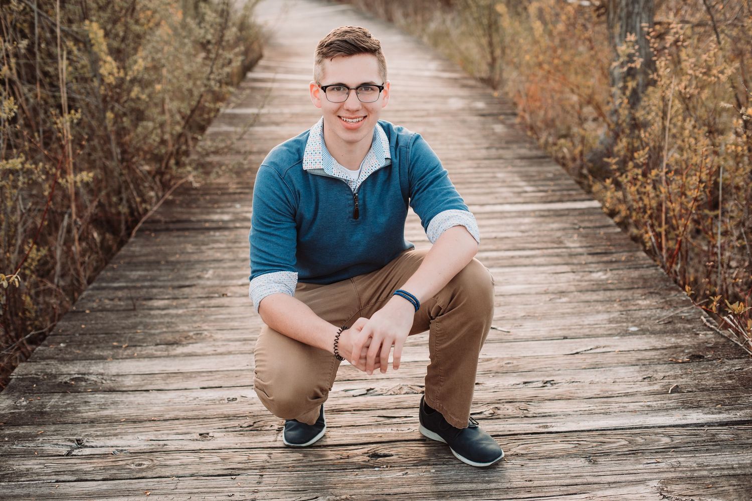 Young man in teal sweater and khakis crouching on a wooden boardwalk, smiling, wearing glasses outdoors.