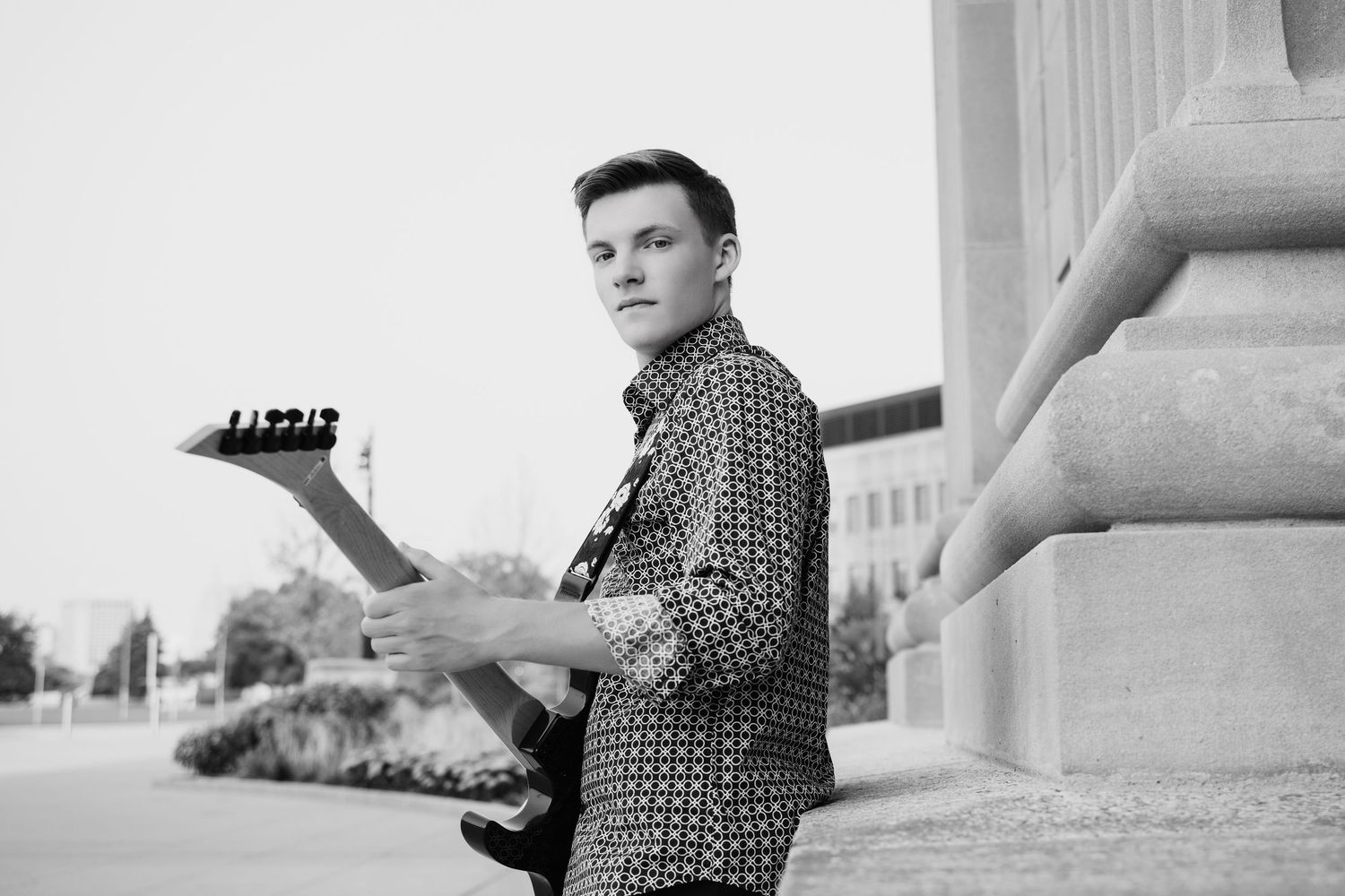 Young male guitarist holding electric guitar outdoors near columns, black and white portrait photo.