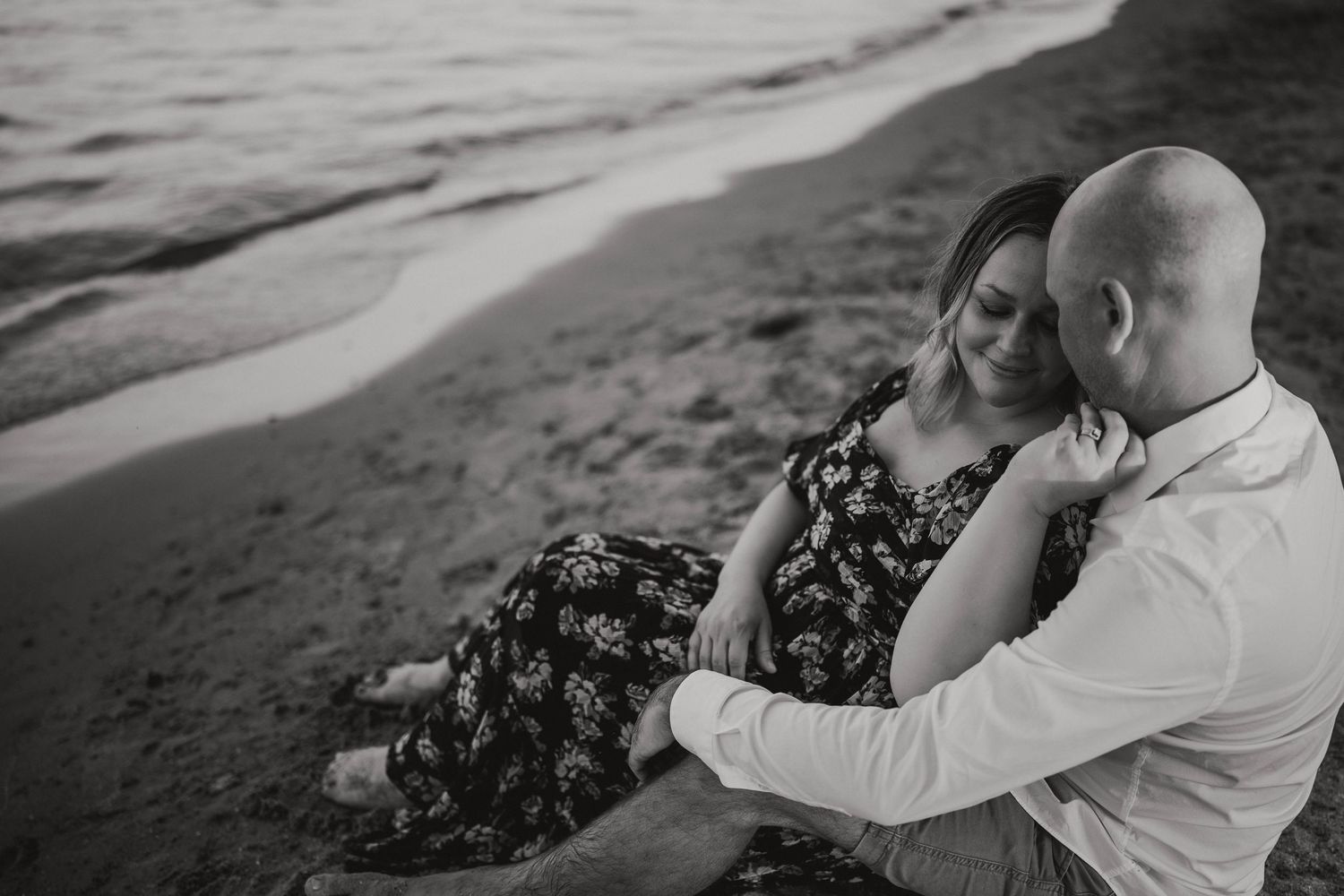 Black and white photo of a couple sitting intimately on a sandy beach at sunset.