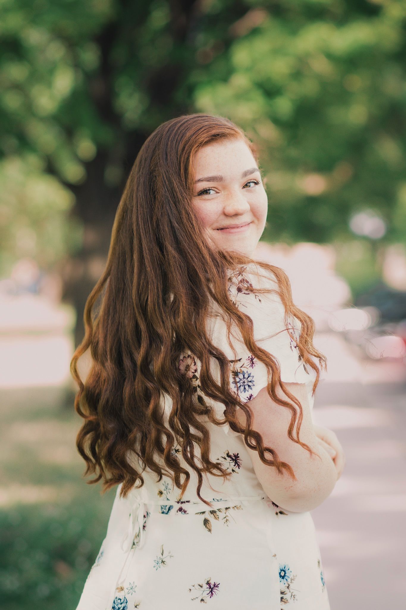 Young woman with long curly auburn hair smiling outdoors in a floral dress with green trees in background.