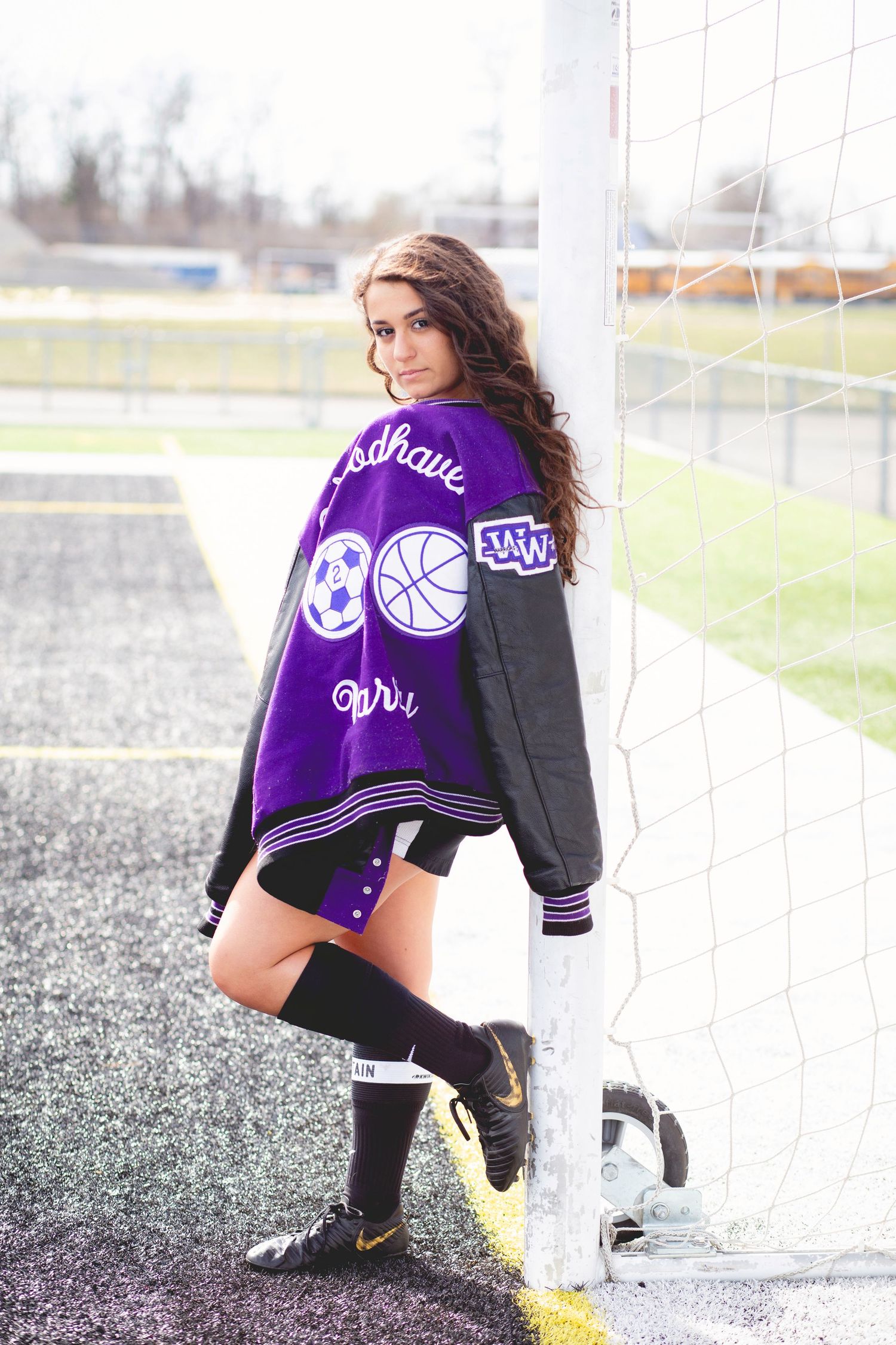 A young female athlete poses in a purple varsity jacket with sports patches near a soccer goal post, wearing knee-high socks.