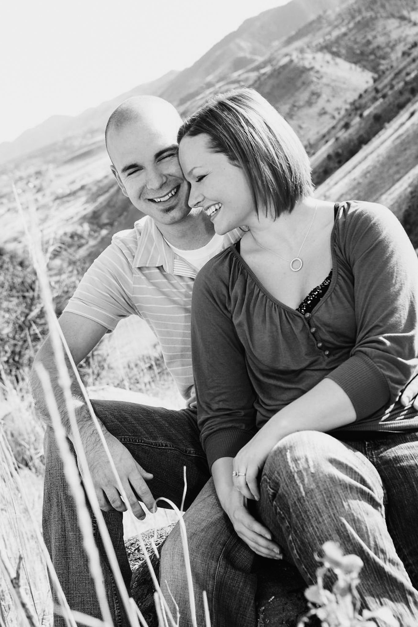 Happy couple sitting together outdoors in a black and white photo, smiling and laughing against a mountain backdrop.