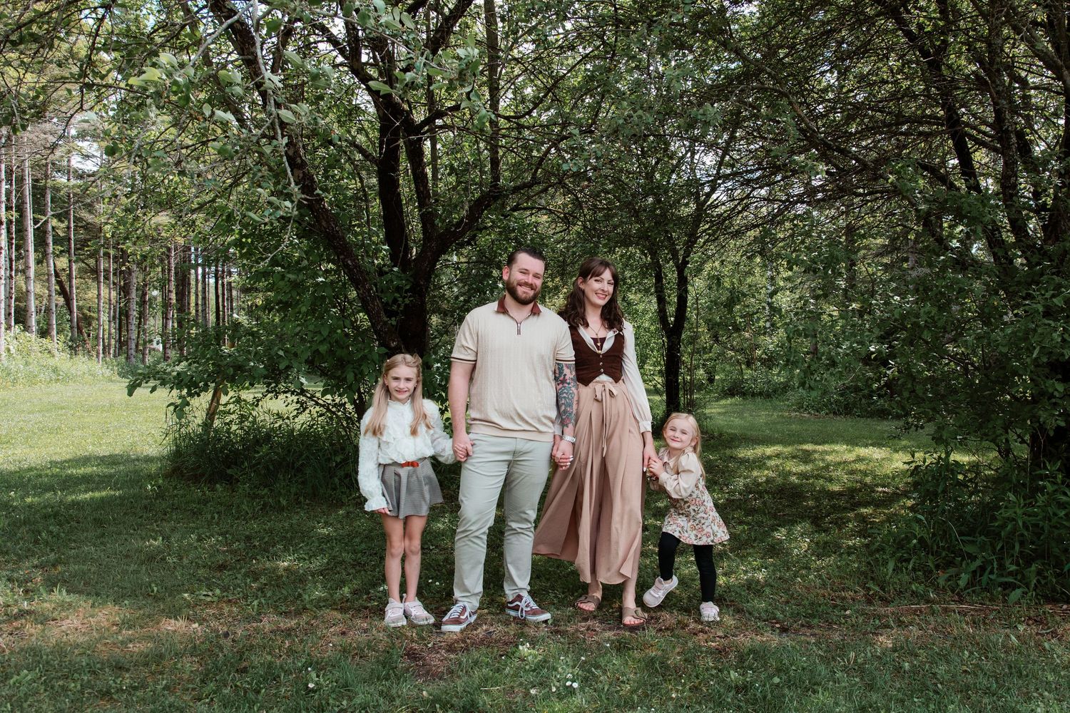Family of four posing outdoors in a lush green park, smiling together on a sunny day.