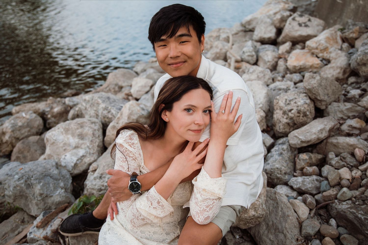 Romantic couple sitting on rocky shore by water, woman in lace dress leaning on man in white shirt.