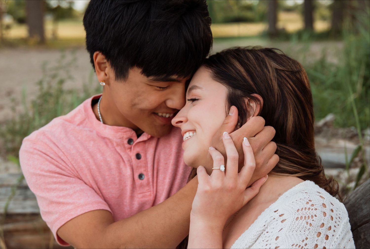 A joyful couple shares a tender moment, the woman displaying an engagement ring while smiling warmly outdoors.