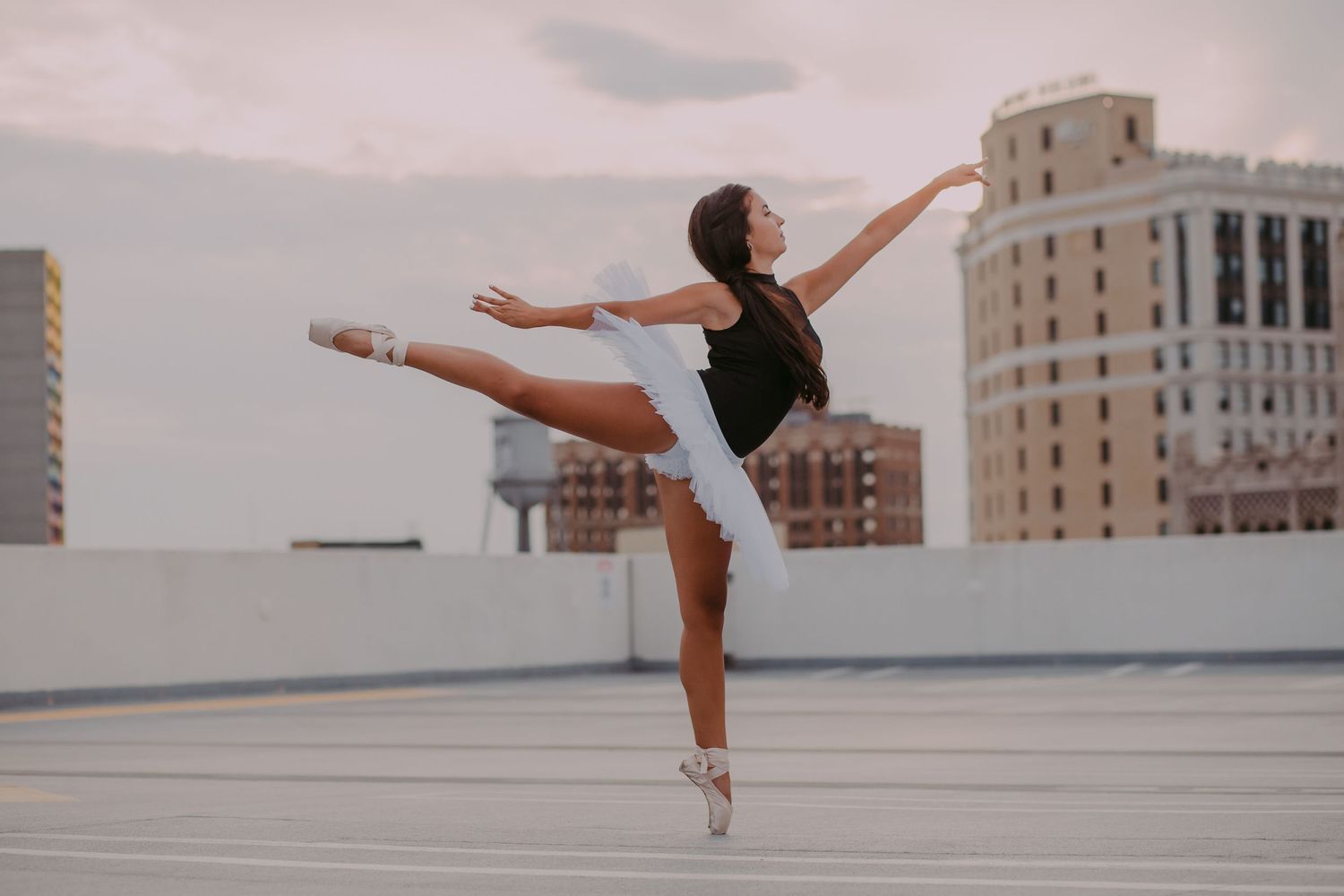 Ballerina in black leotard and white tutu performs an arabesque on a rooftop with city buildings behind her.