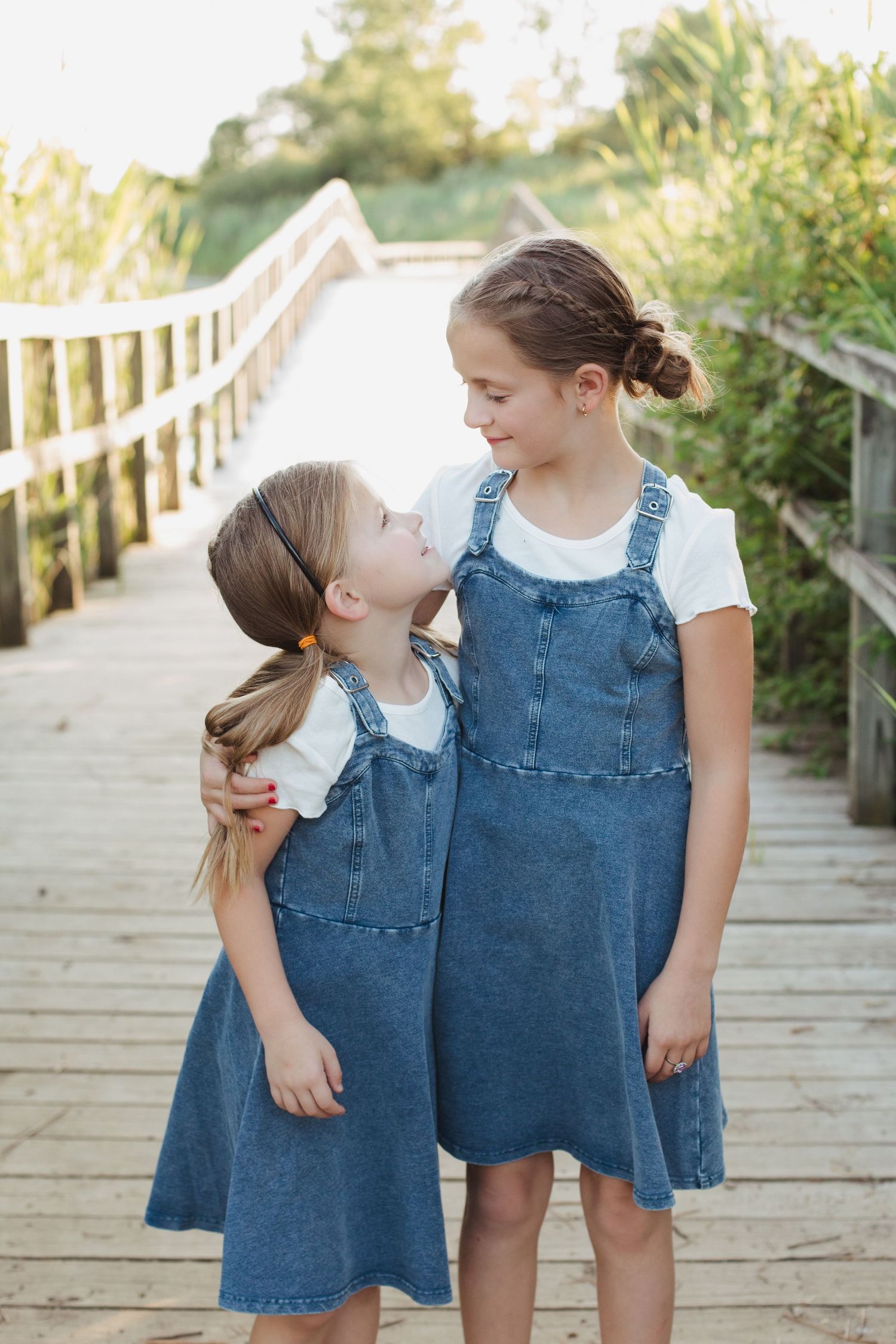 Two young sisters in matching denim jumpers smile at each other on a sunny wooden bridge outdoors.
