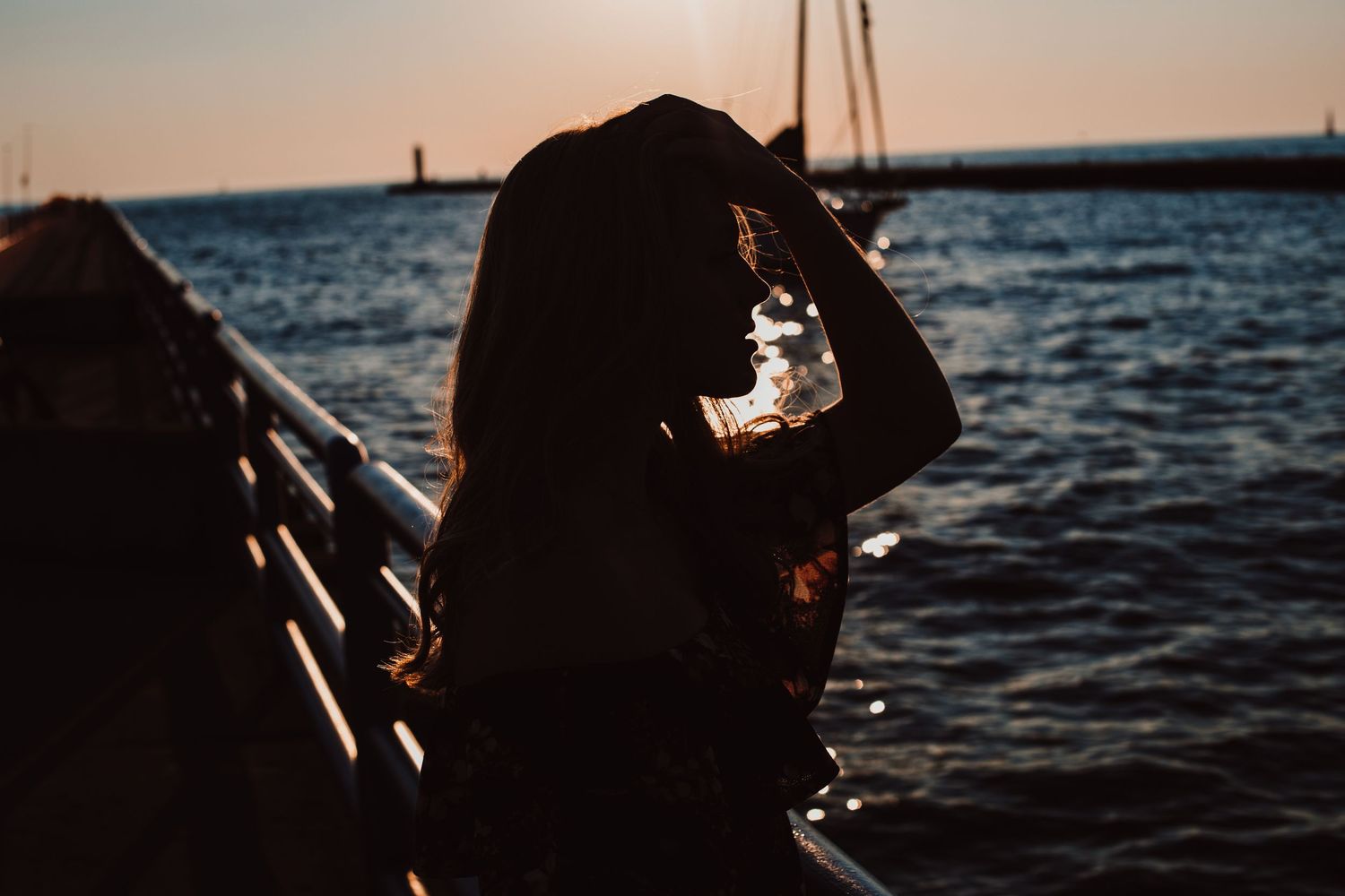 Silhouette of woman holding water bottle by waterfront railing at golden sunset.