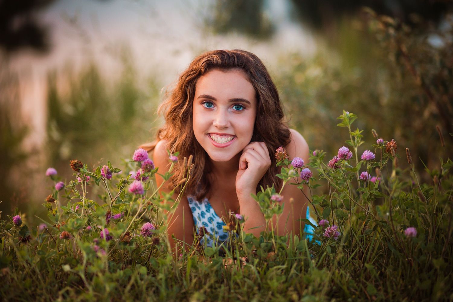 Smiling young woman lying among purple wildflowers in sun-drenched meadow wearing blue top.
