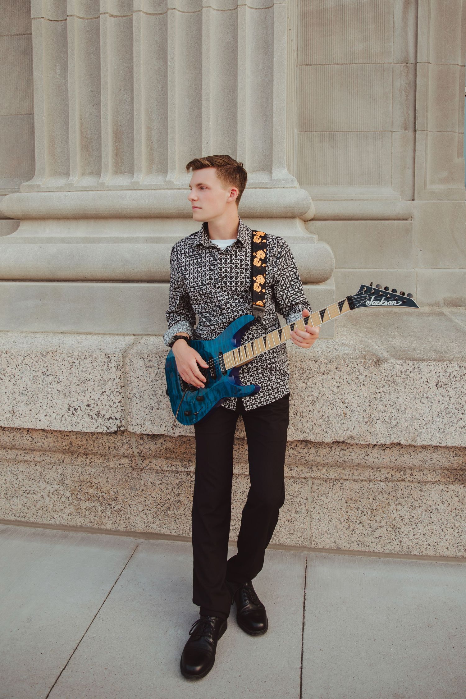 Young man holding a blue electric guitar with floral strap while leaning against a stone building column.