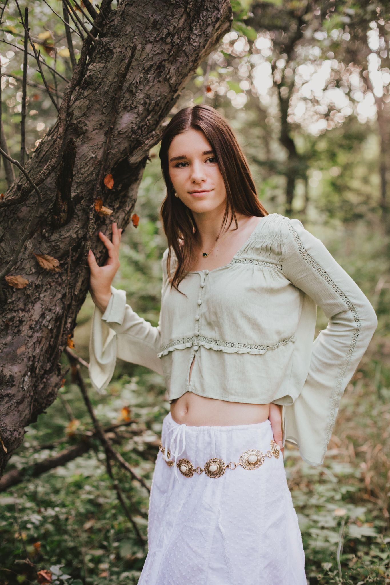 Young woman in bohemian crop top and white skirt poses by a tree in a lush forest setting.