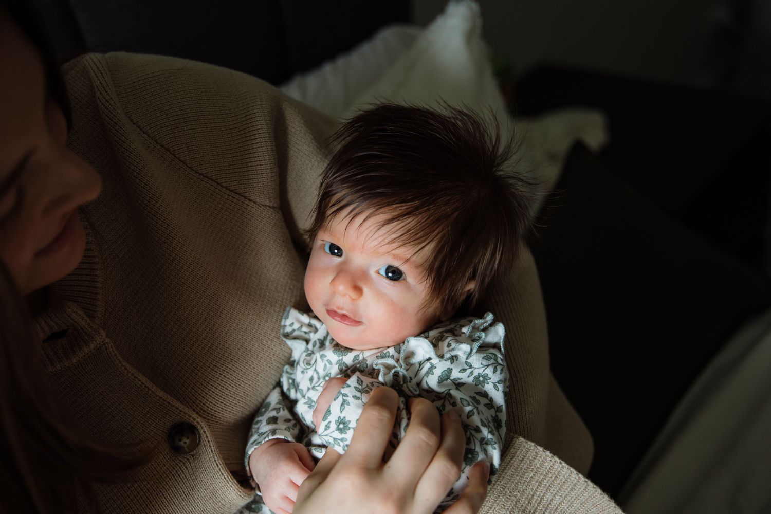 Newborn baby with dark hair in floral onesie being held by parent in warm light.